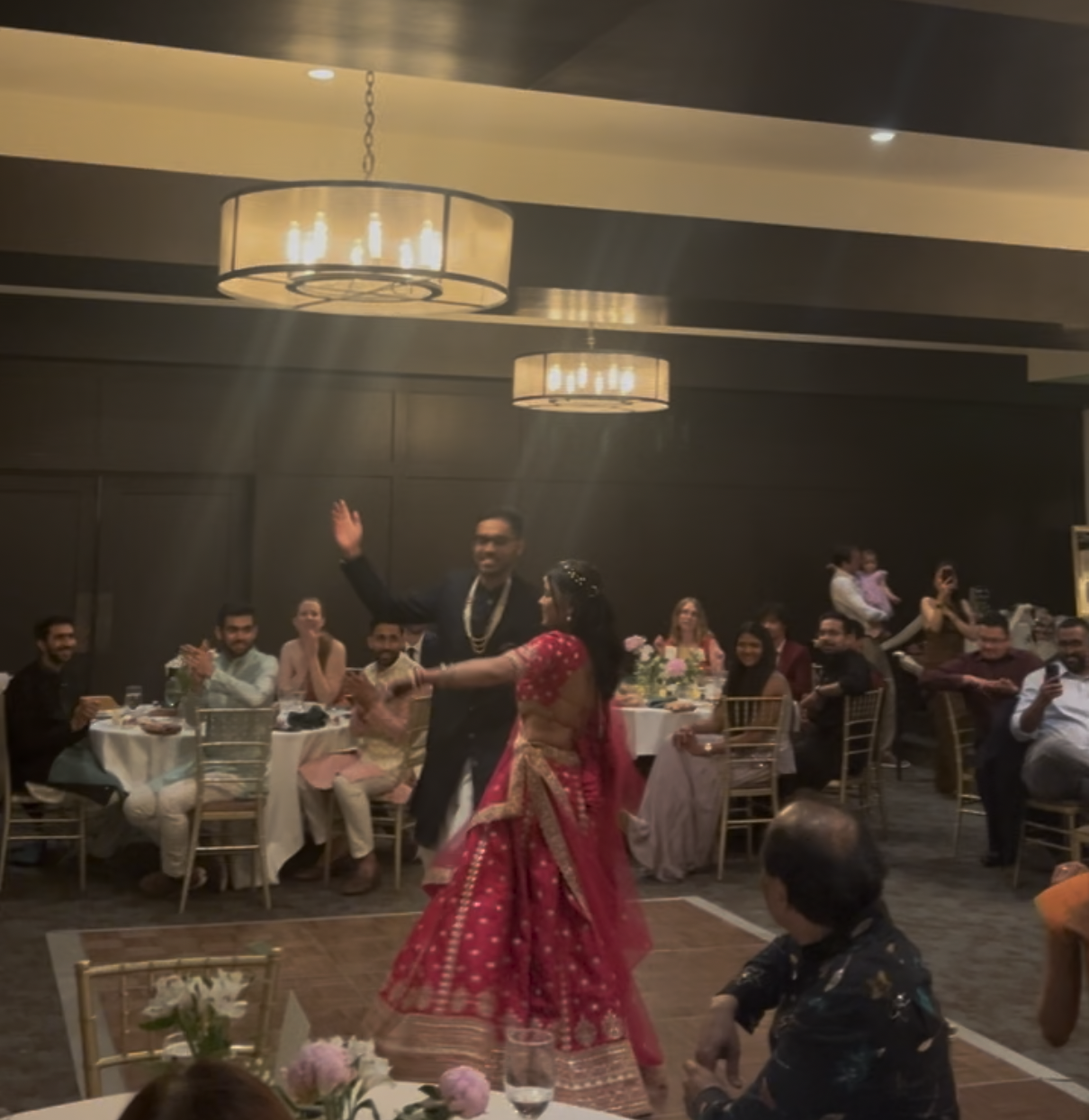 People celebrating at a wedding reception, with a couple dancing on the dance floor in traditional Indian attire, surrounded by guests seated at decorated tables.