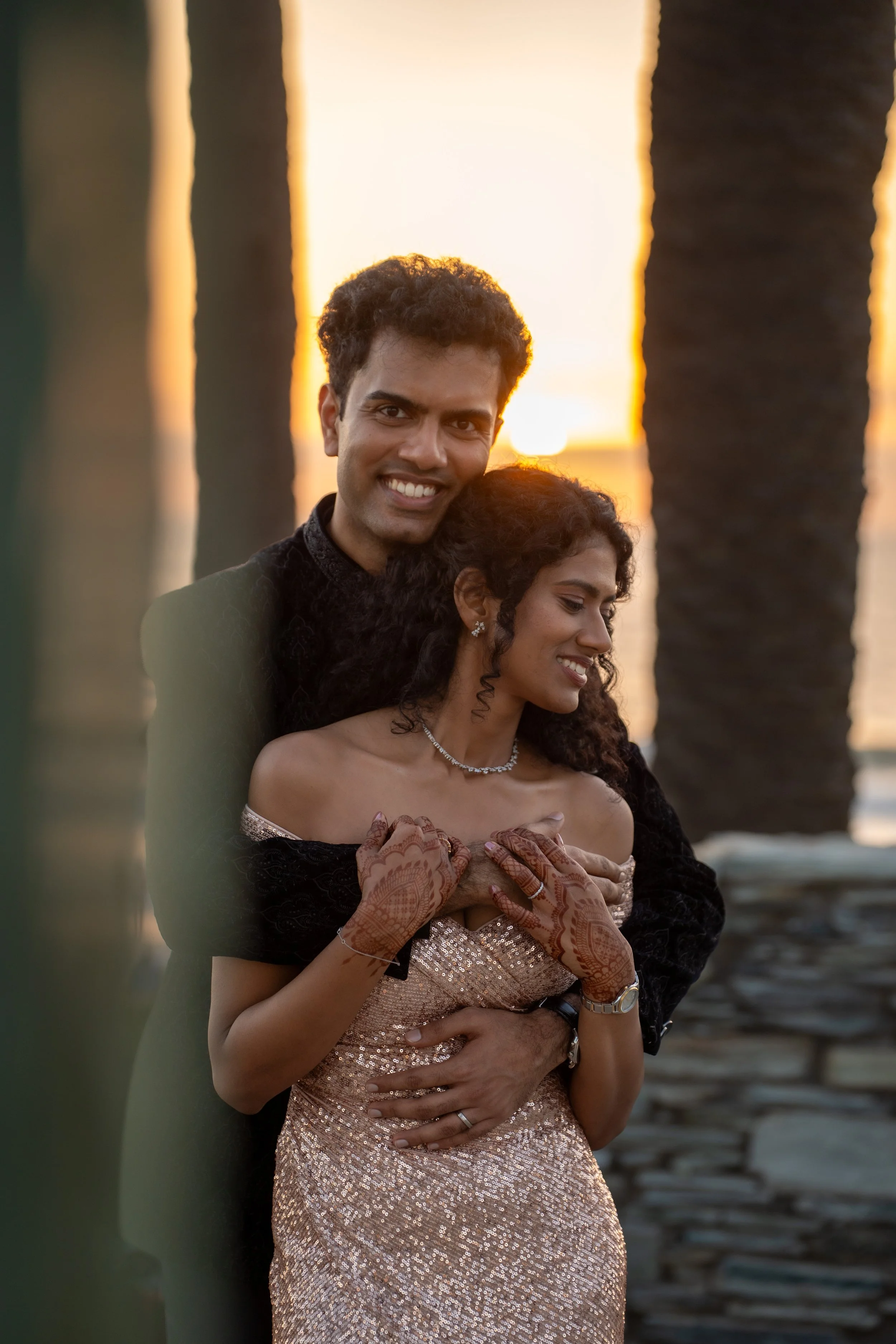 A smiling man and woman embrace on a beach at sunset, with palm trees in the background. The woman wears a sparkling gold dress and traditional henna on her hands, and the man wears a black outfit. They appear happy and intimate.
