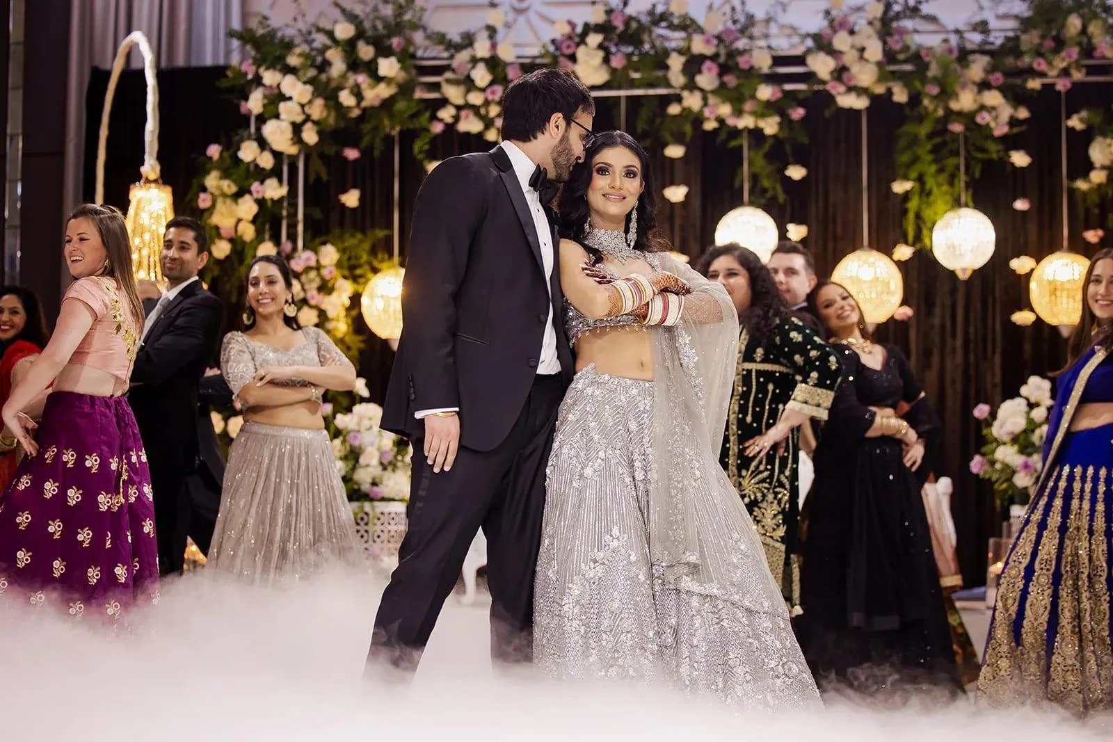 A bride and groom dancing at their wedding reception, surrounded by friends and family in festive traditional attire, with floral and lantern decorations in the background.
