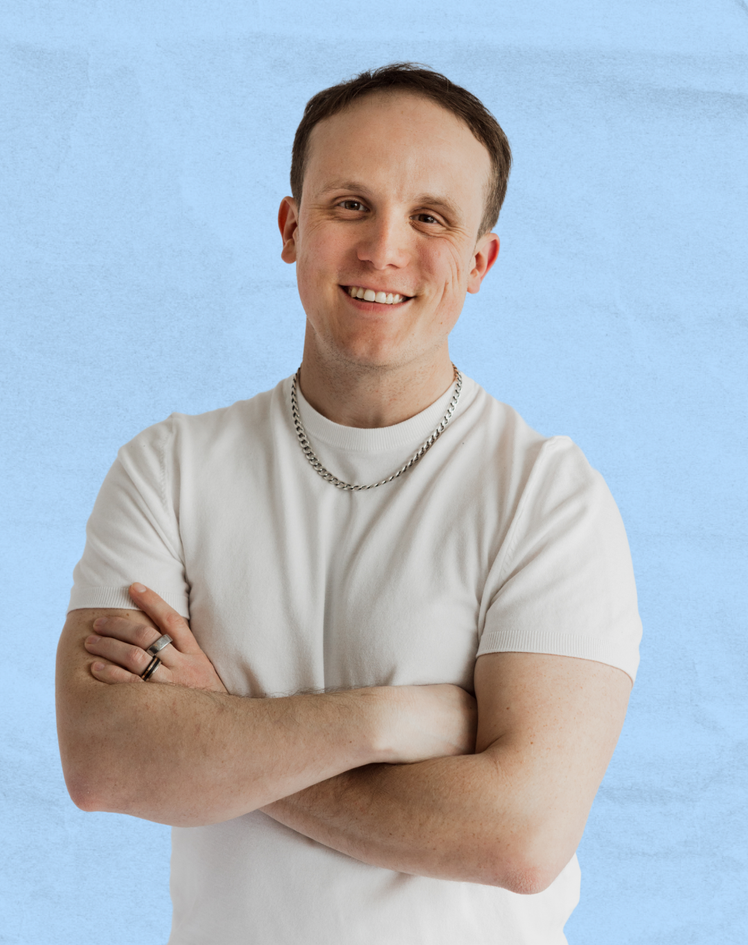 A young man with short brown hair smiling, wearing a white t-shirt and silver jewelry, standing with crossed arms against a light blue background.