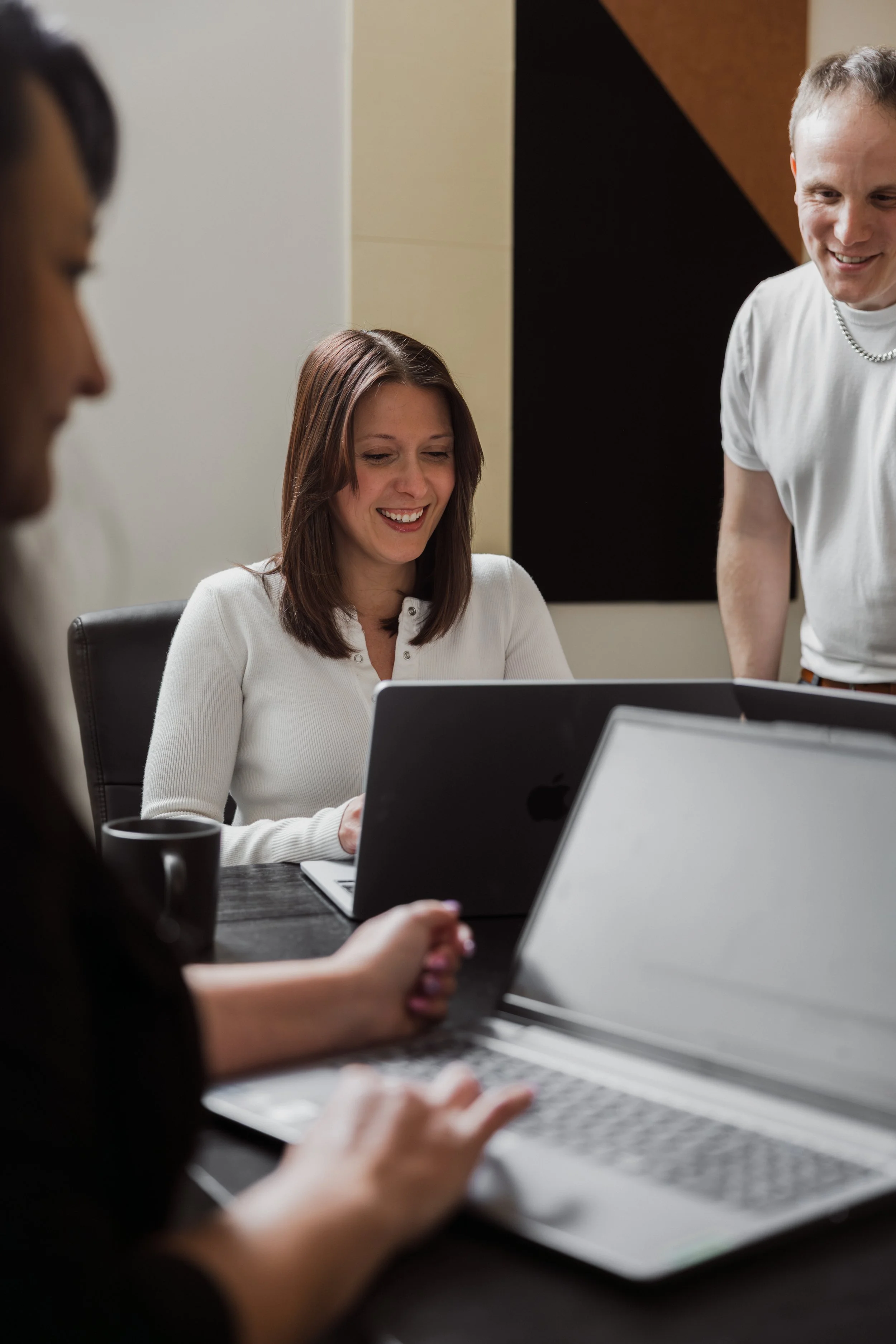 Young woman in white shirt smiling at laptop in an office, two other people partially visible using laptops in the foreground.