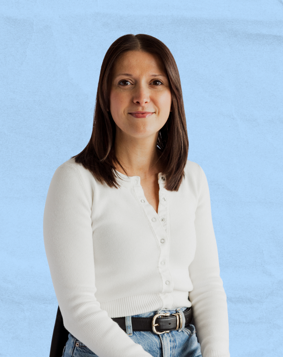A woman with shoulder-length brown hair, wearing a white button-up shirt and blue jeans, standing with a light blue textured background.