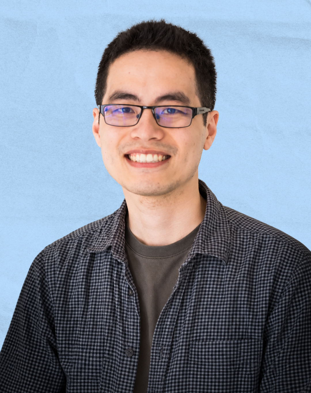 Young man with dark hair and glasses smiling against a light blue background.