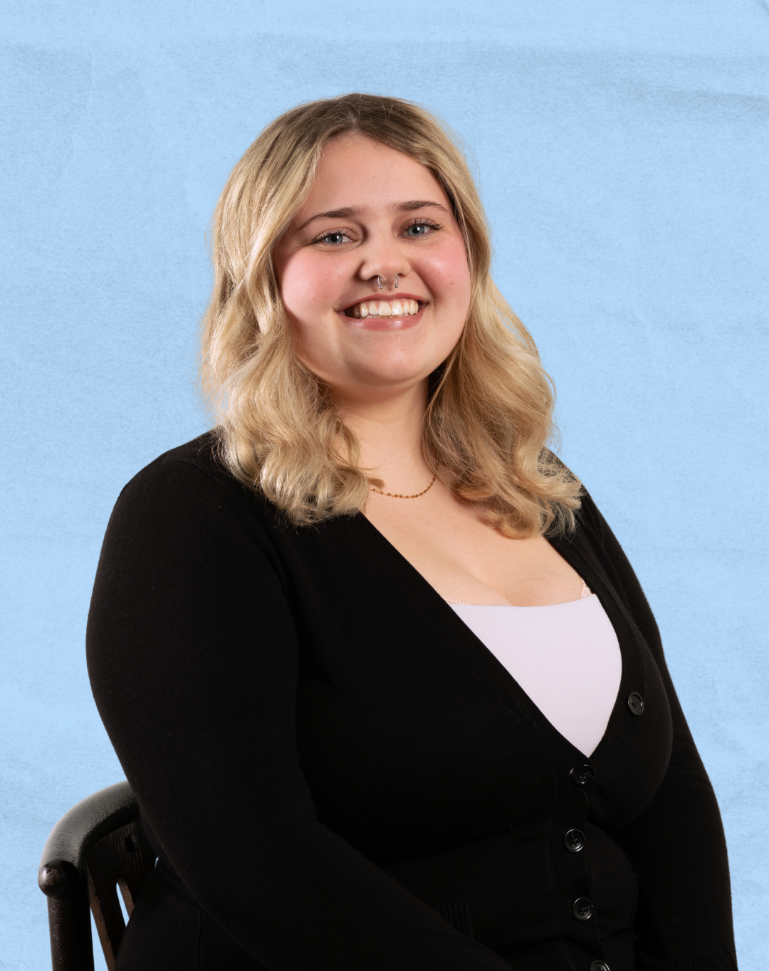 A young woman with blonde hair, smiling, wearing a black sweater over a white top, sitting on a black chair against a textured light blue background.