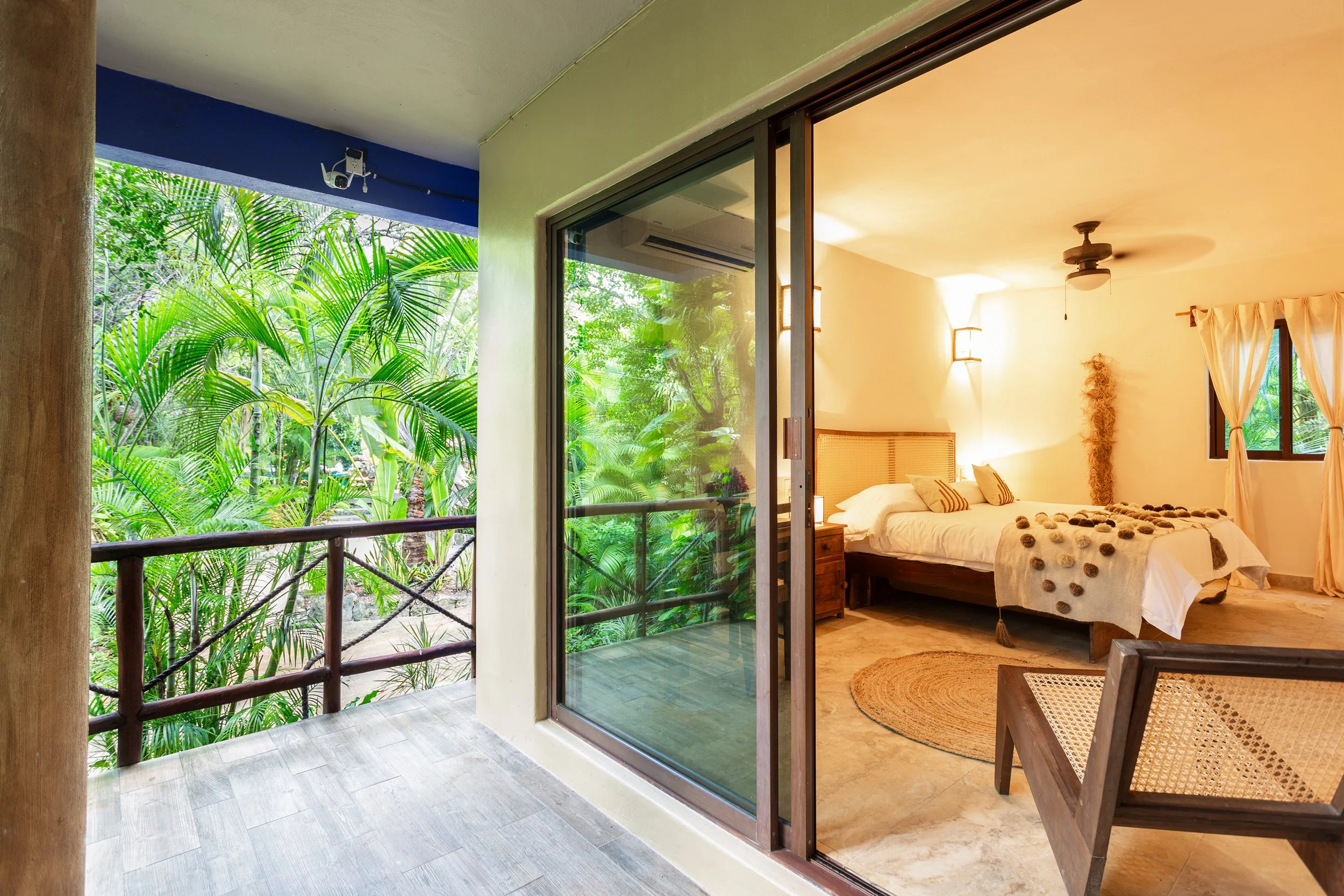 A bedroom with beige walls and a large bed with white linens and decorative pillows. There's a wooden nightstand, a round woven rug, and a ceiling fan. The room has a window with cream curtains and a partially open sliding door leading to a balcony w