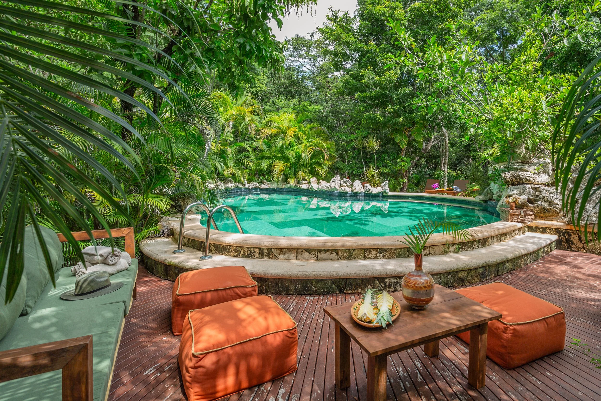 A backyard swimming pool surrounded by lush green tropical plants and trees, with outdoor furniture including a wooden table, cushions, and a vase on a wooden deck.