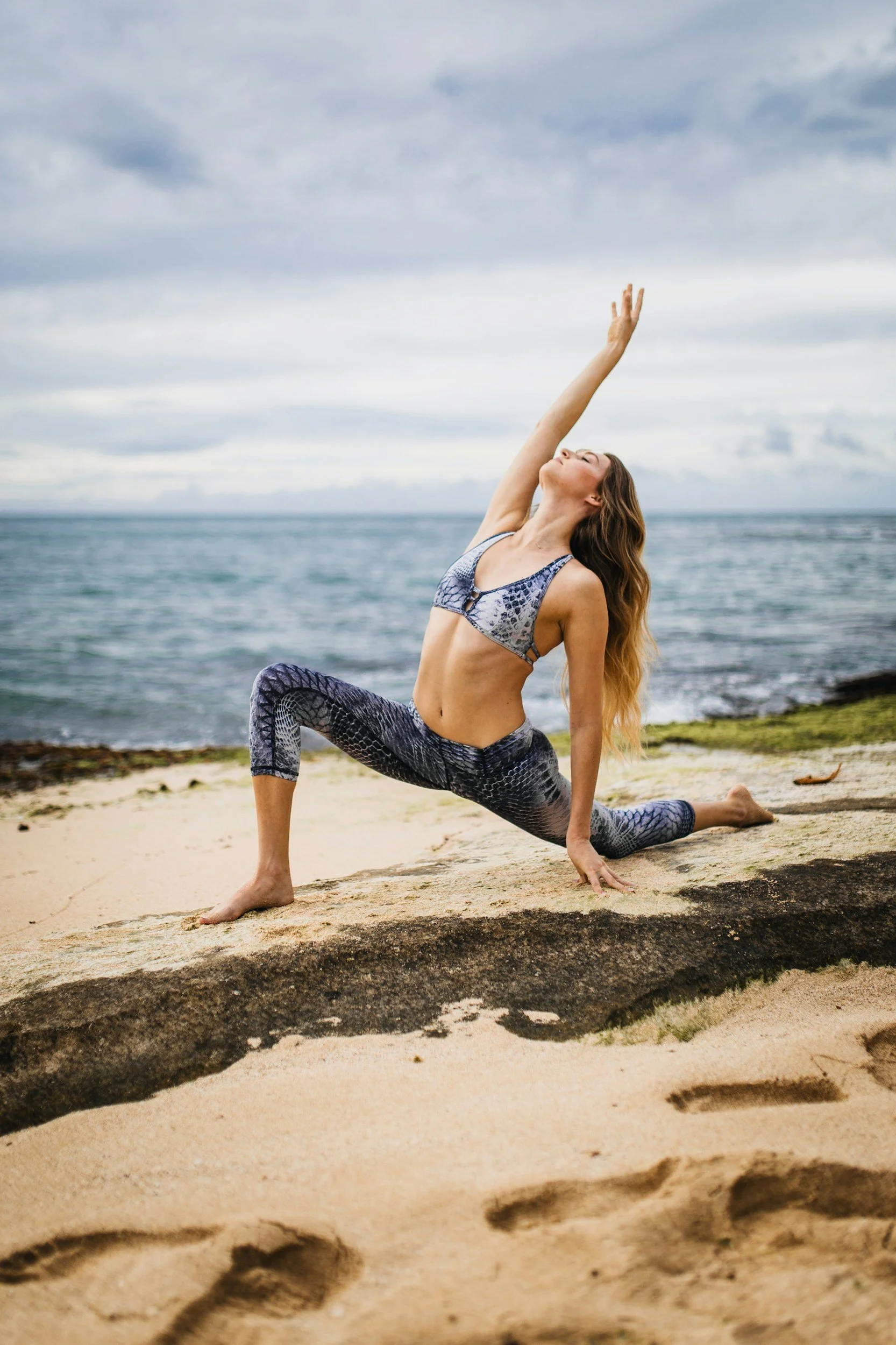 A woman practicing yoga on a beach, performing a low lunge pose with one arm extended upward and the other hand on the ground, wearing a snakeskin-patterned workout outfit.