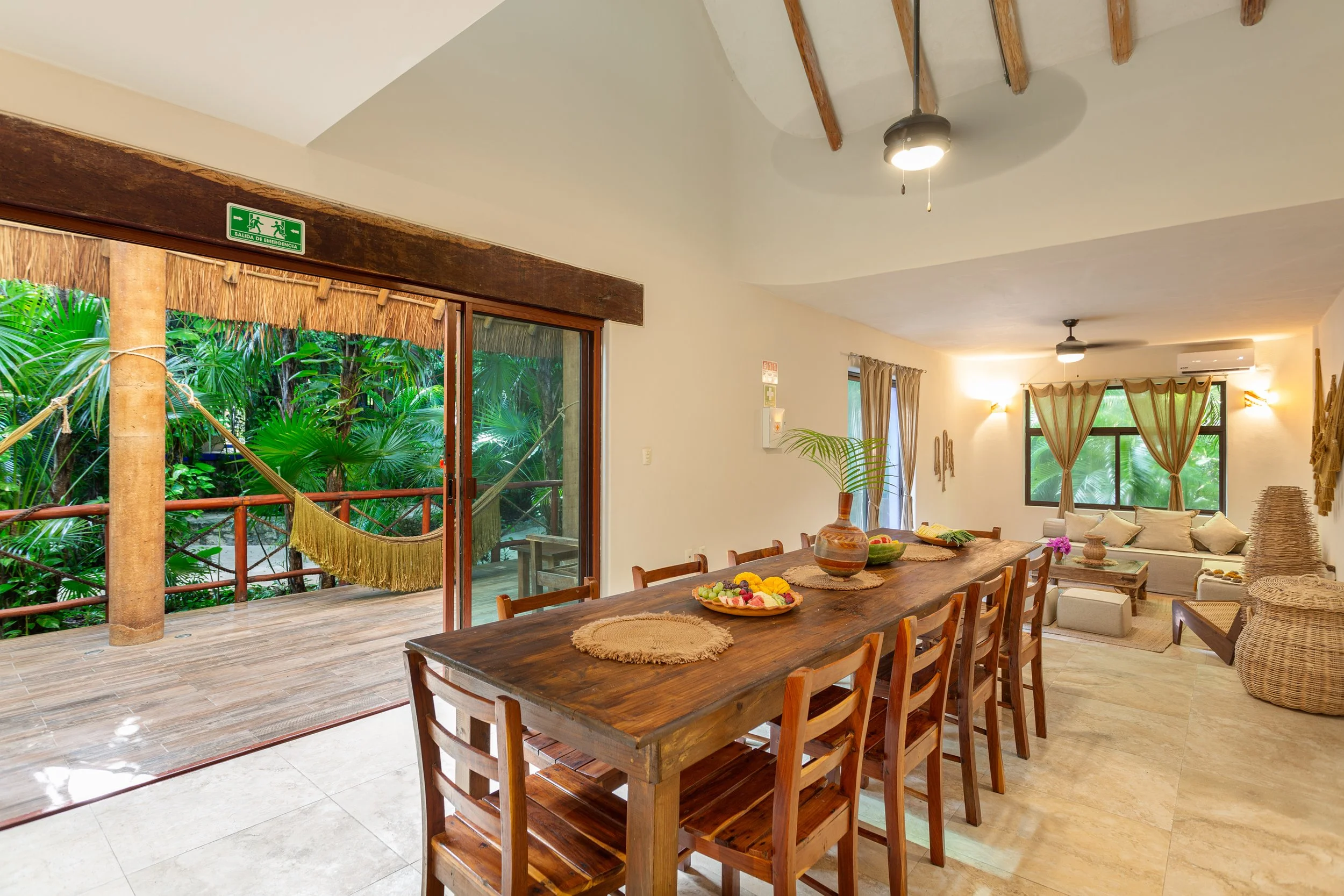 Open-plan dining and living area with a large wooden dining table, chairs, and a sitting area with a sofa. Large sliding glass doors open to a patio with greenery. Decor includes vases, bowls, and wall art, with natural light and tropical plants outs