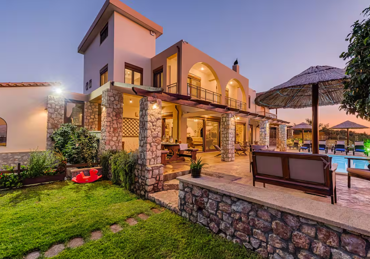 Luxury house with stone accents, outdoor seating area, swimming pool, and thatched umbrellas at dusk.