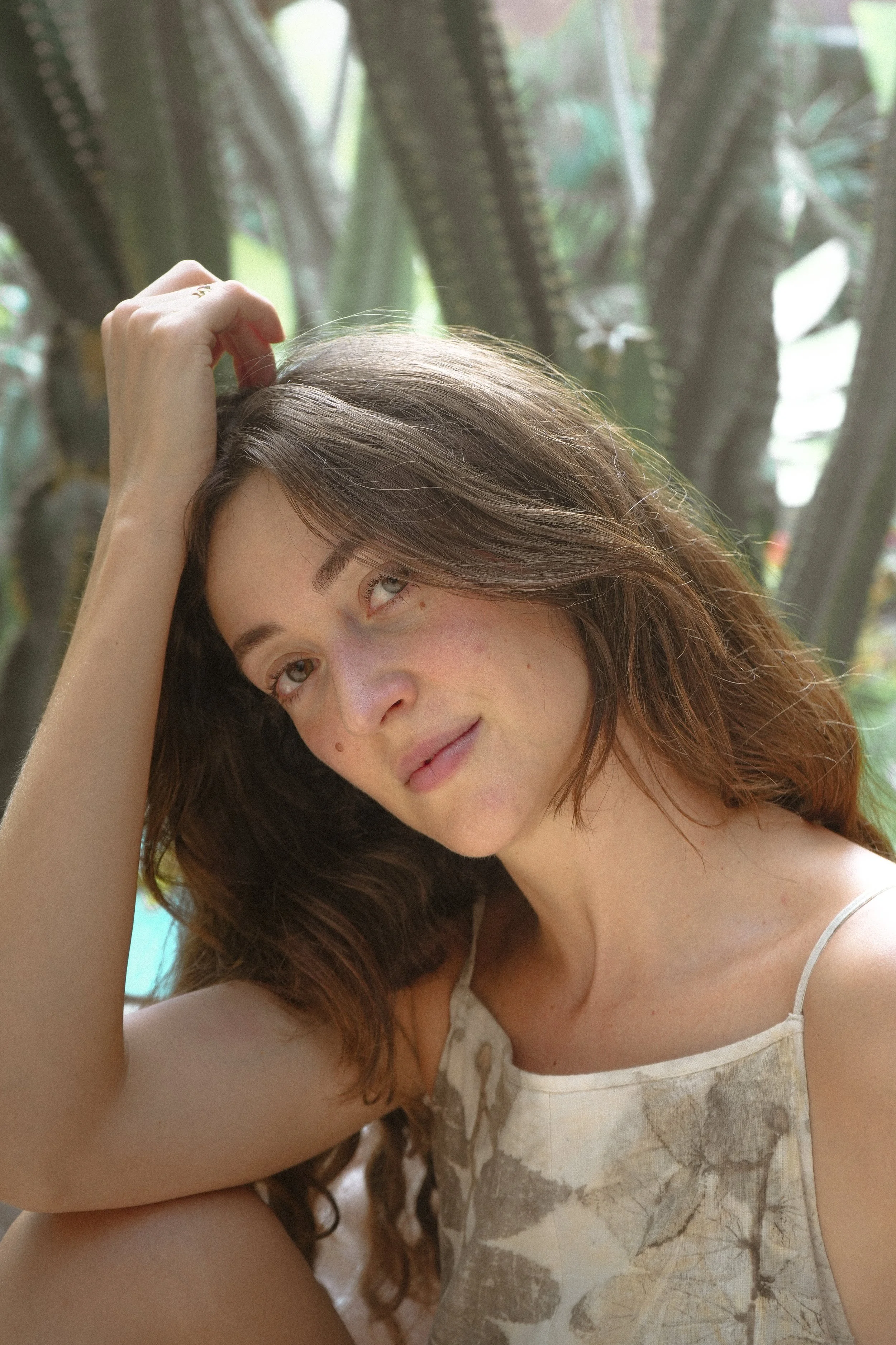 A young woman with brown hair and a patterned sleeveless top, sitting outdoors in front of green plants, gazing softly at the camera with her head resting on her hand.