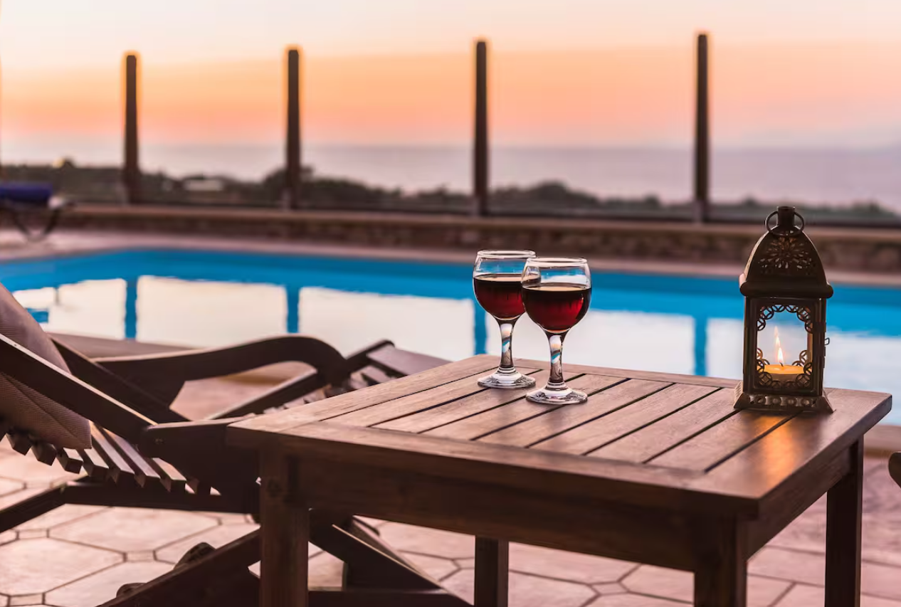 A poolside patio at sunset with two glasses of red wine, a lantern with a candle, and lounge chairs.