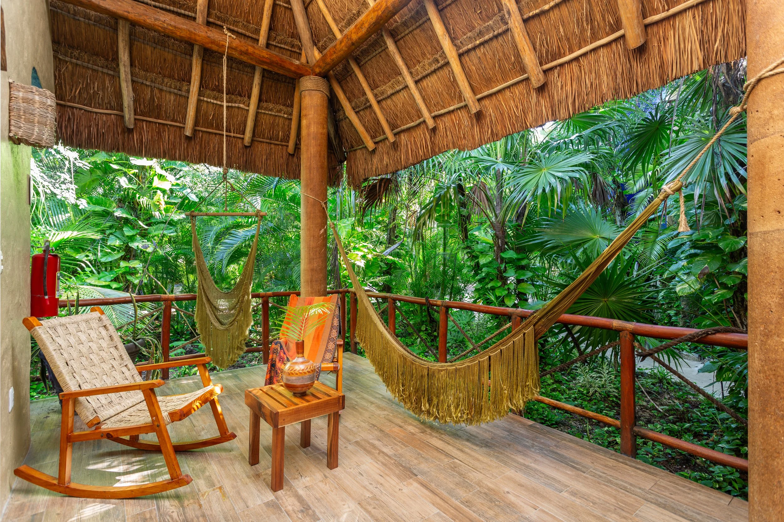 A porch area with wooden rocking chair, small table with a decorative vase, hammock, and lush green tropical plants in the background.