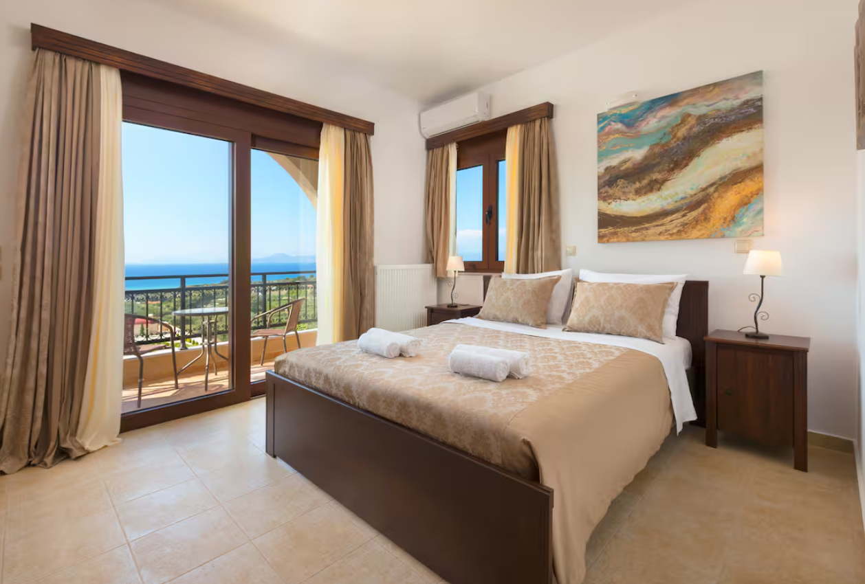 A hotel room with a large bed, two rolled towels on it, and a balcony with outdoor furniture, overlooking a scenic view of the ocean and greenery.