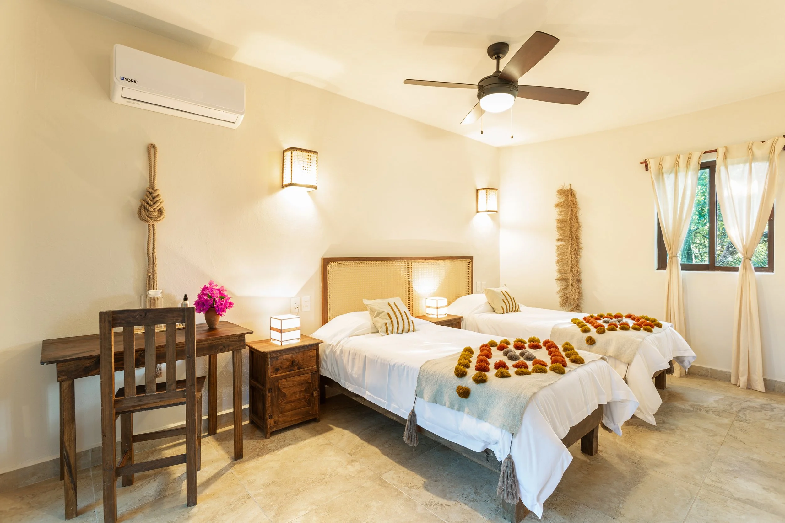 A bedroom with two beds, decorated with flowers spelling out 'LOVE', a wooden desk with a chair, a nightstand with a lamp, wall sconces, a ceiling fan, and a window with cream curtains.
