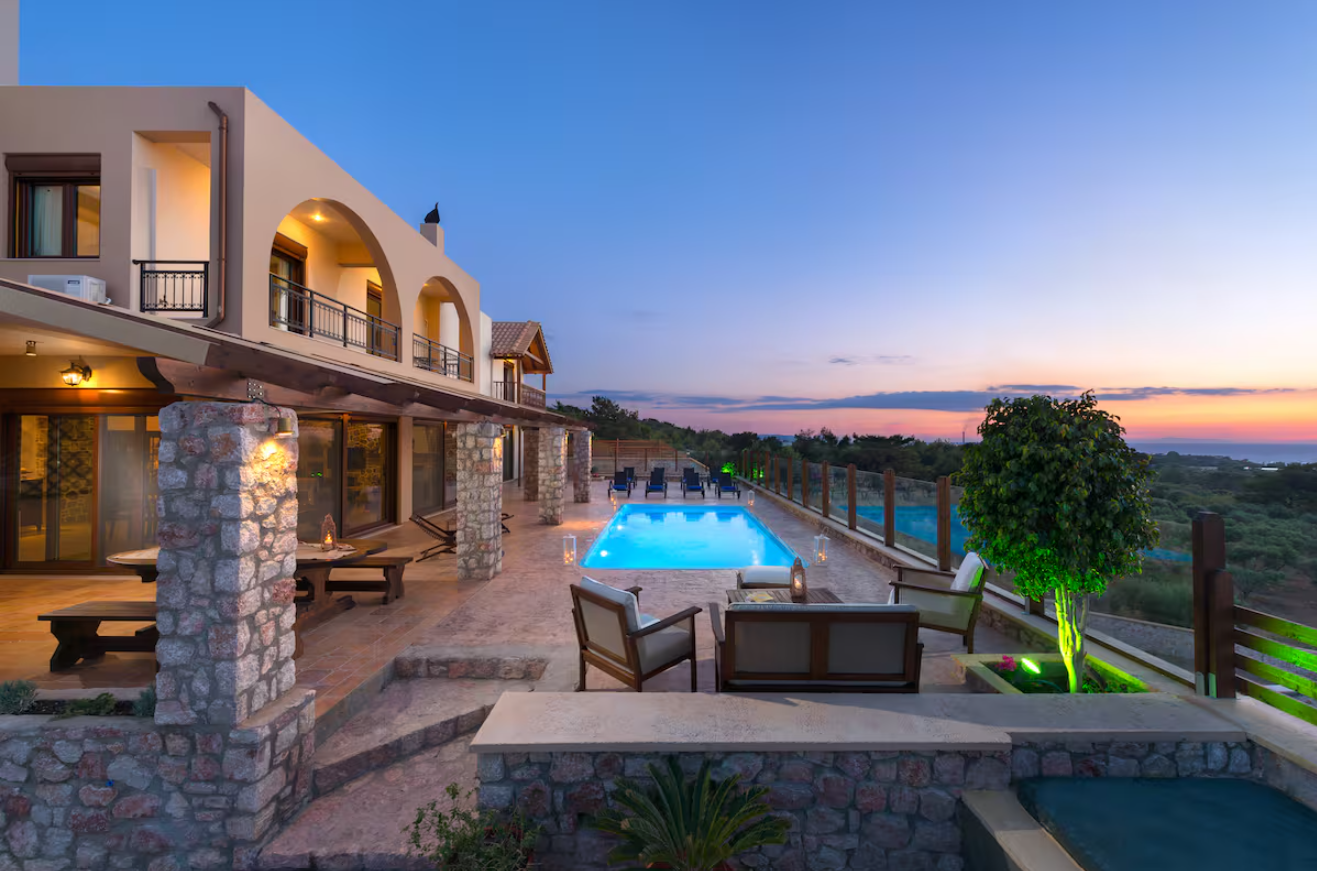 Luxury villa with a swimming pool, outdoor seating, and scenic sunset view with greenery and ocean in the distance.