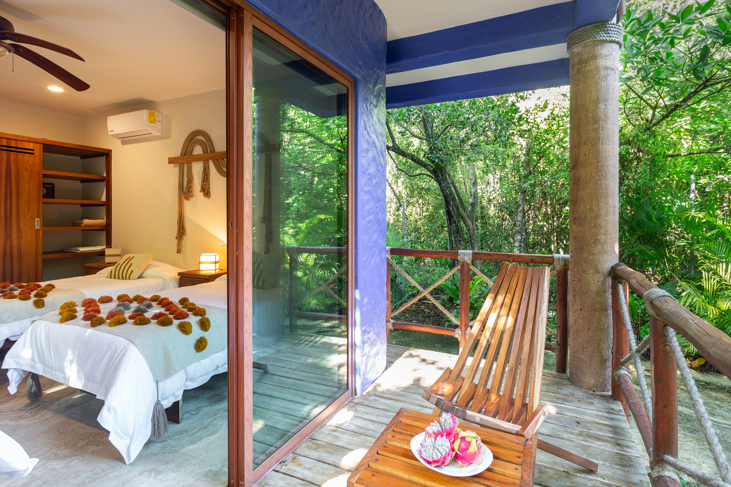 Hotel room with two beds decorated with colorful pom-poms, wooden furniture, and a balcony overlooking lush green trees, with a plate of carved dragon fruit on a wooden chair.