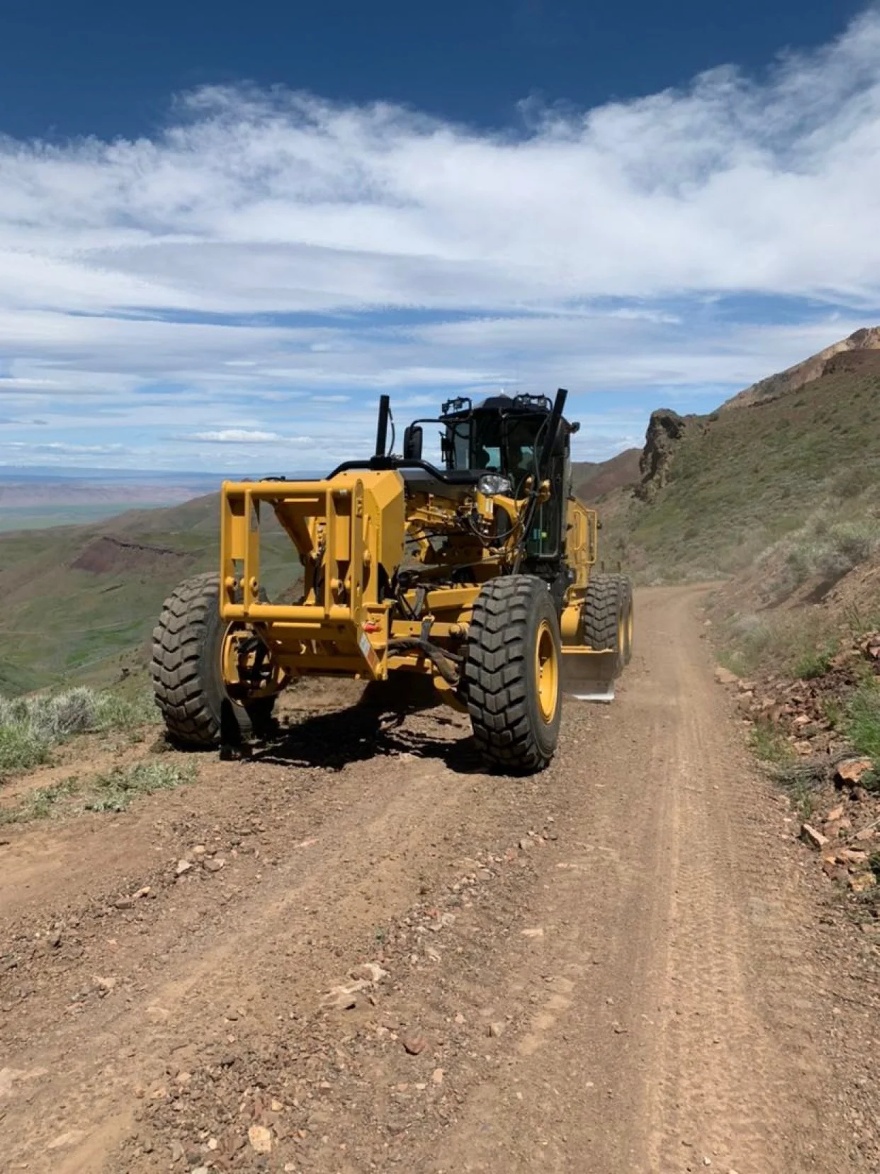 Caterpillar motor grader parked on a graded dirt mountain road above a high desert valley