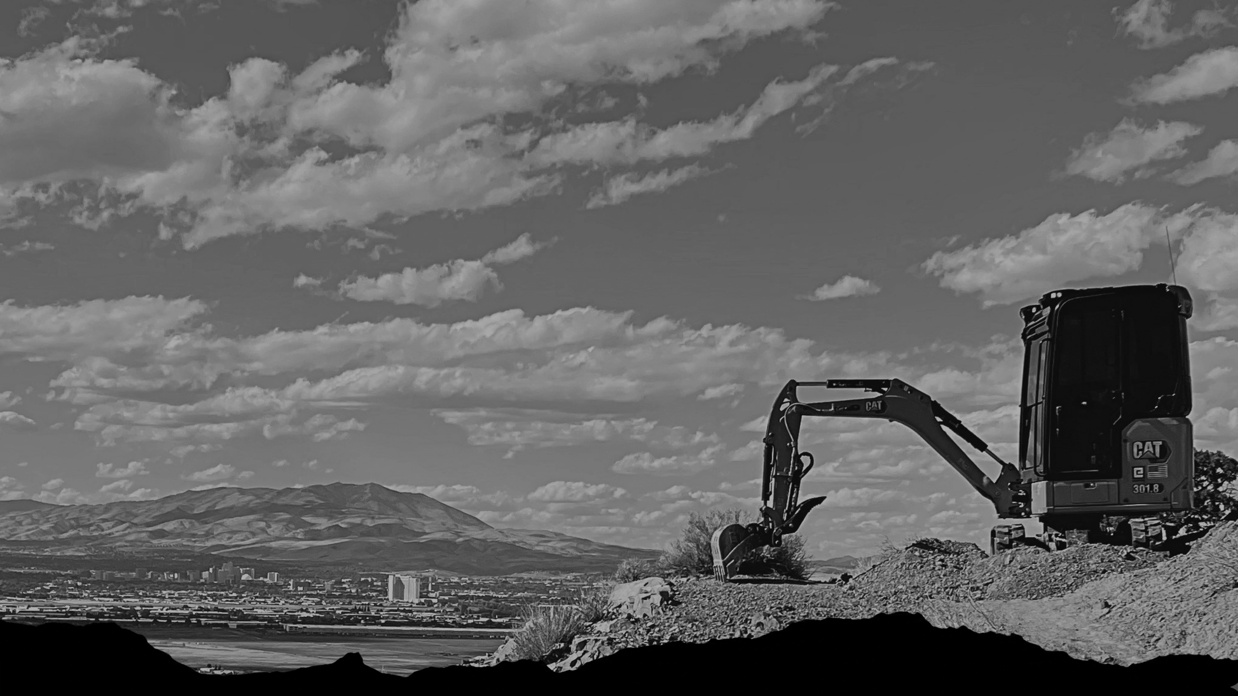 A black and white photograph of a CAT mini excavator parked on a hillside overlooking Reno and distant mountains under a cloudy sky.