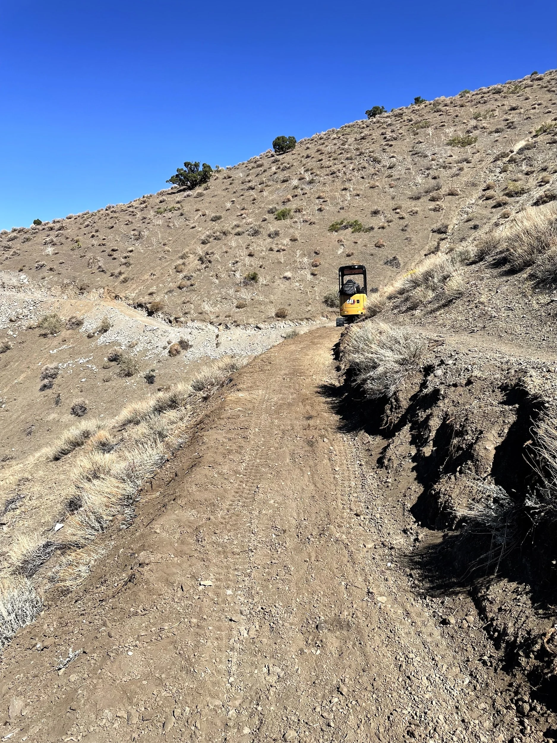 Natural surface trail up a brushy hillside with an excavator in the distance