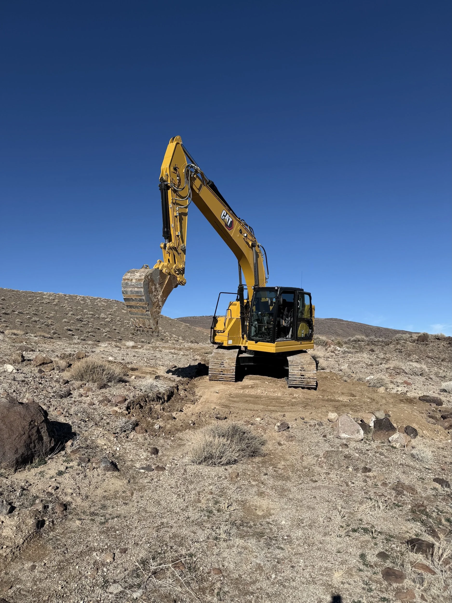 A yellow excavator working on a rocky, dry landscape under a clear blue sky.