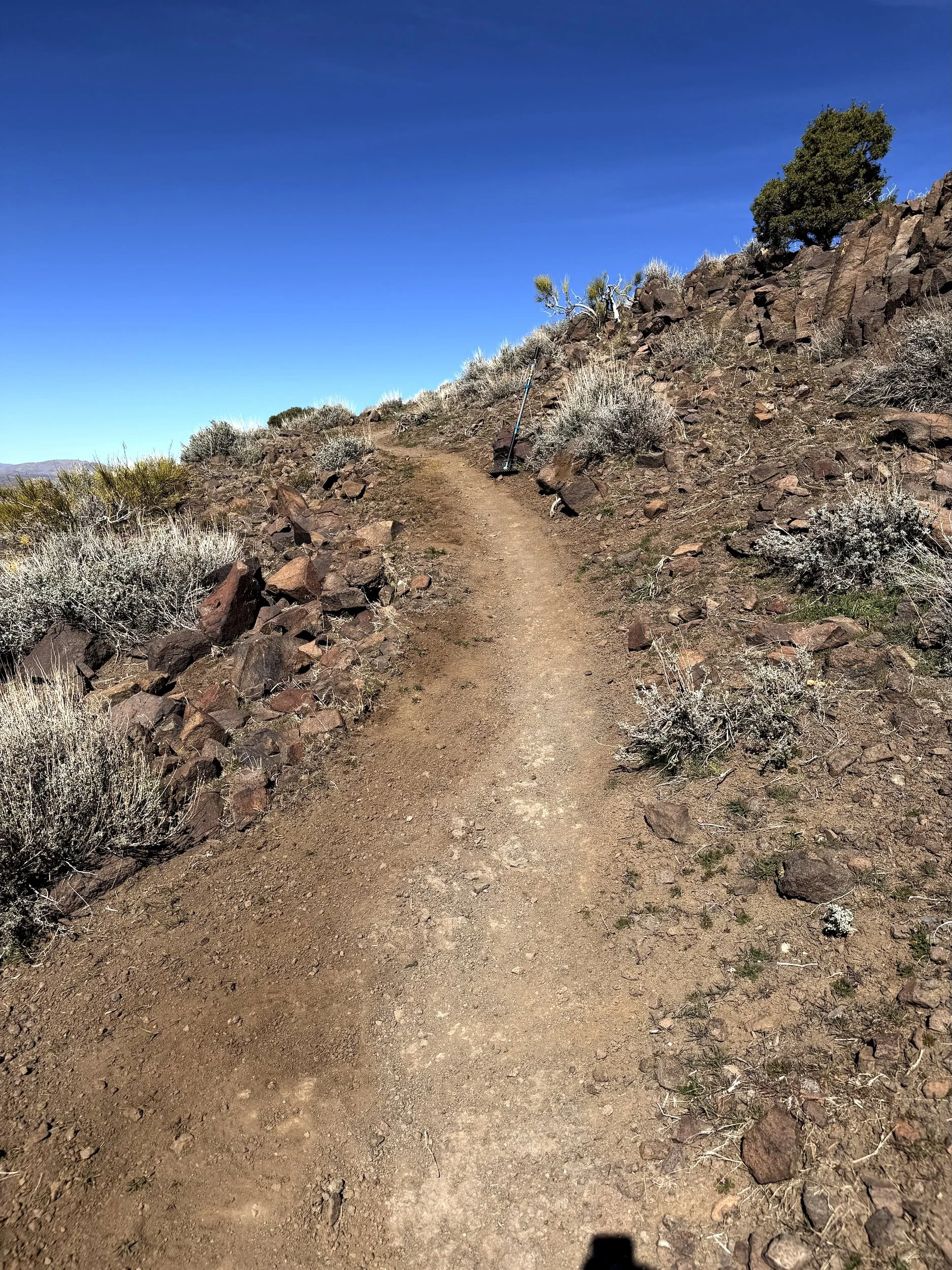 Newly built natural surface trail climbing a rocky sagebrush hillside, with a hand tool resting against the rock work.