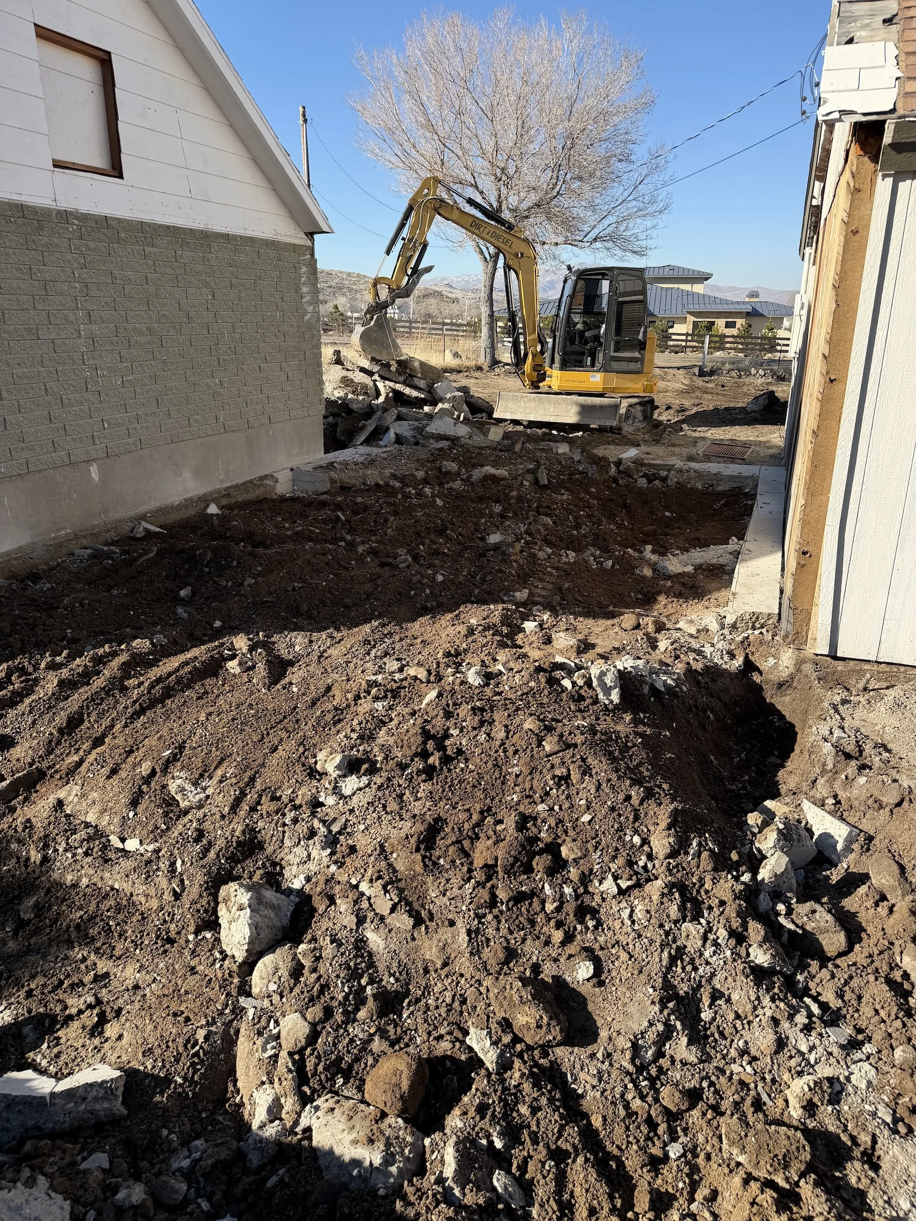 Excavator clearing and grading a dirt site next to a house