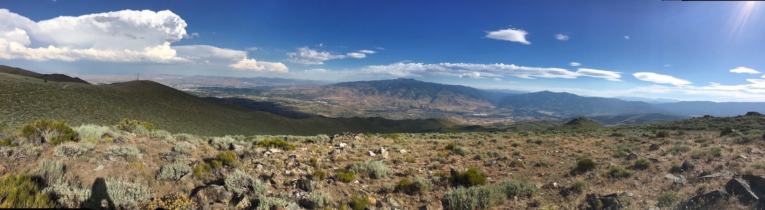 Panoramic view of a mountainous landscape with a mix of green and brown hills, under a partly cloudy blue sky with bright sunlight.