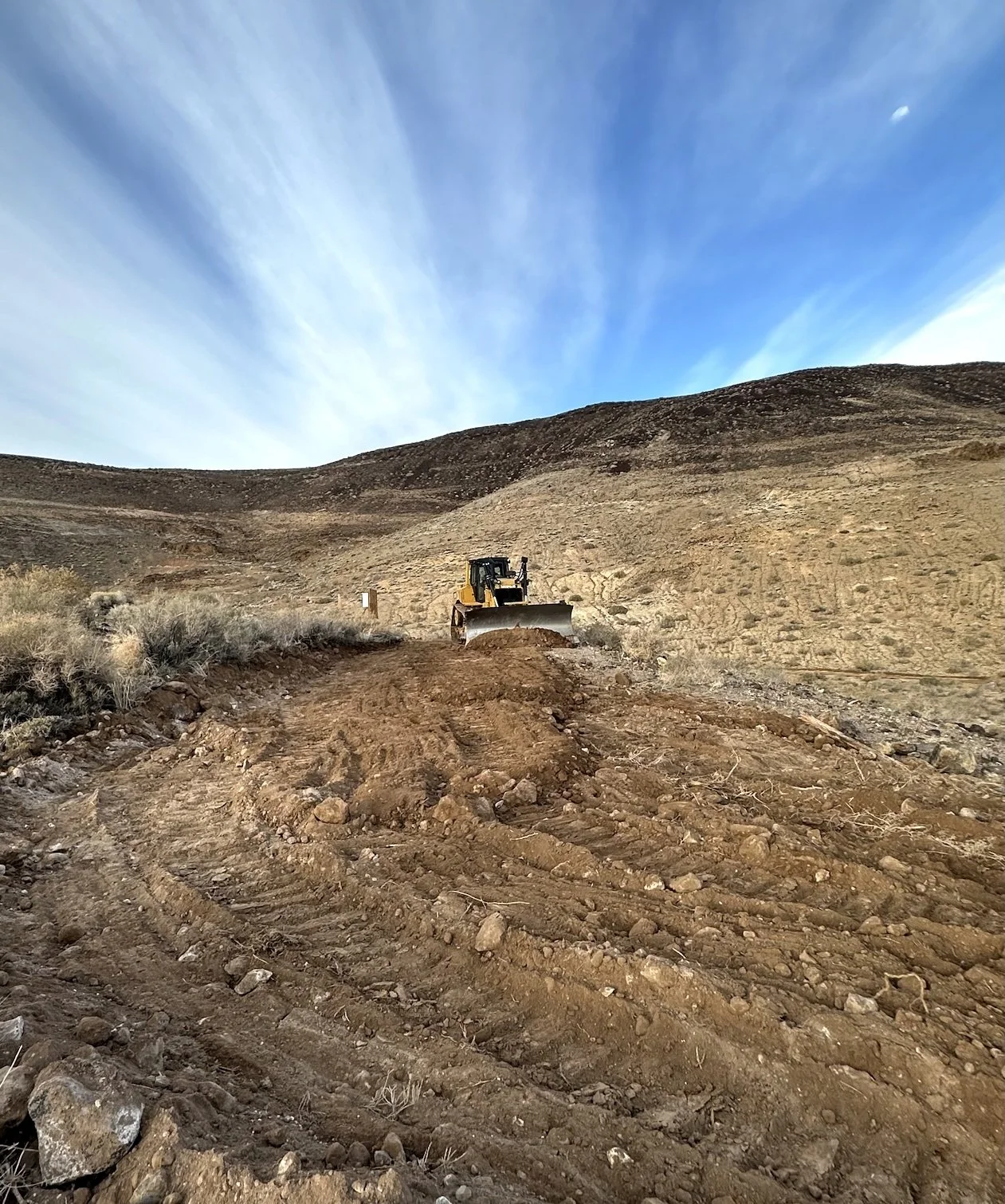 Heavy equipment grading a rural dirt road on a sagebrush hillside