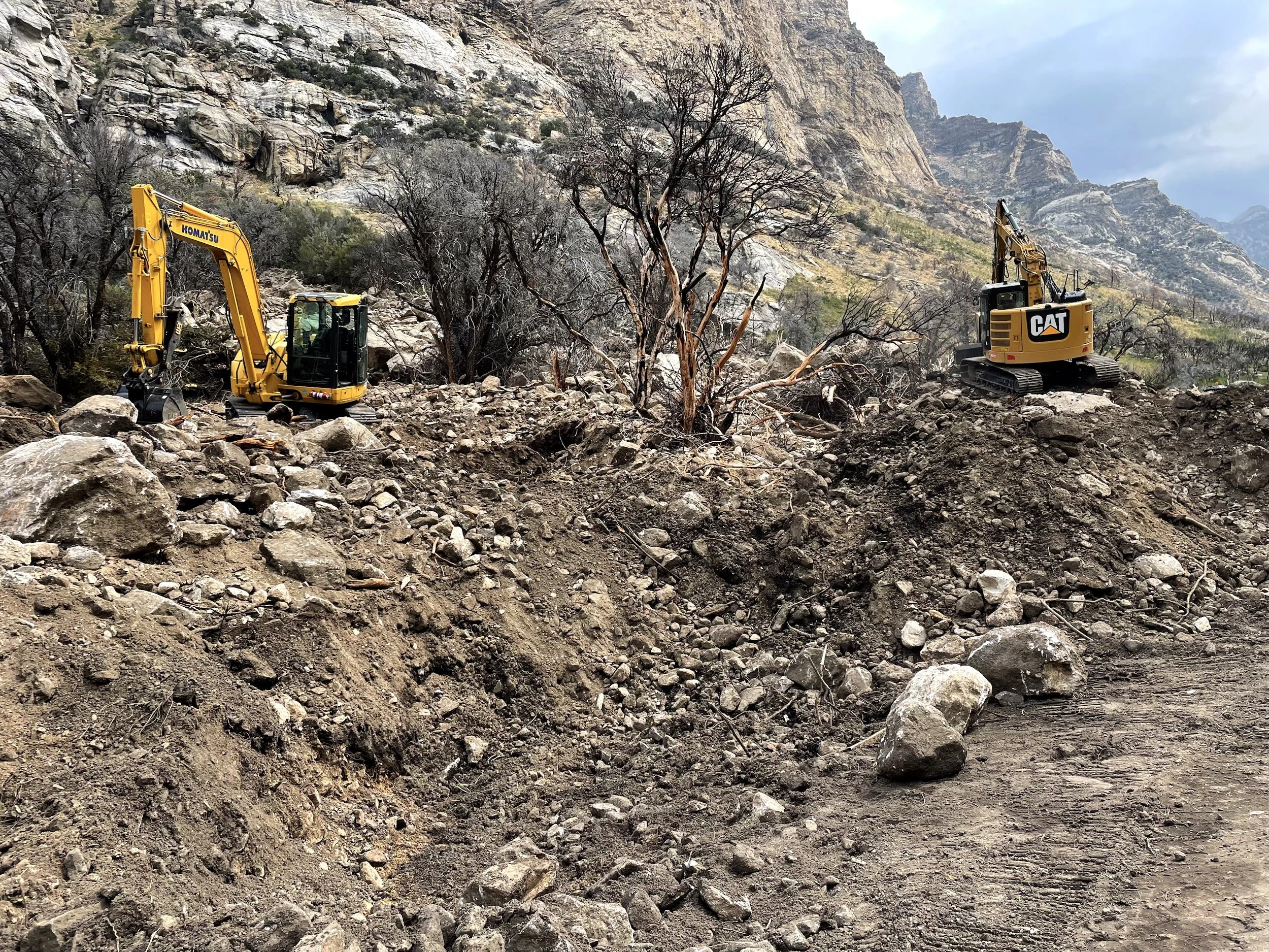 Excavator clearing a steep rocky hillside scattered with boulders and burned trees