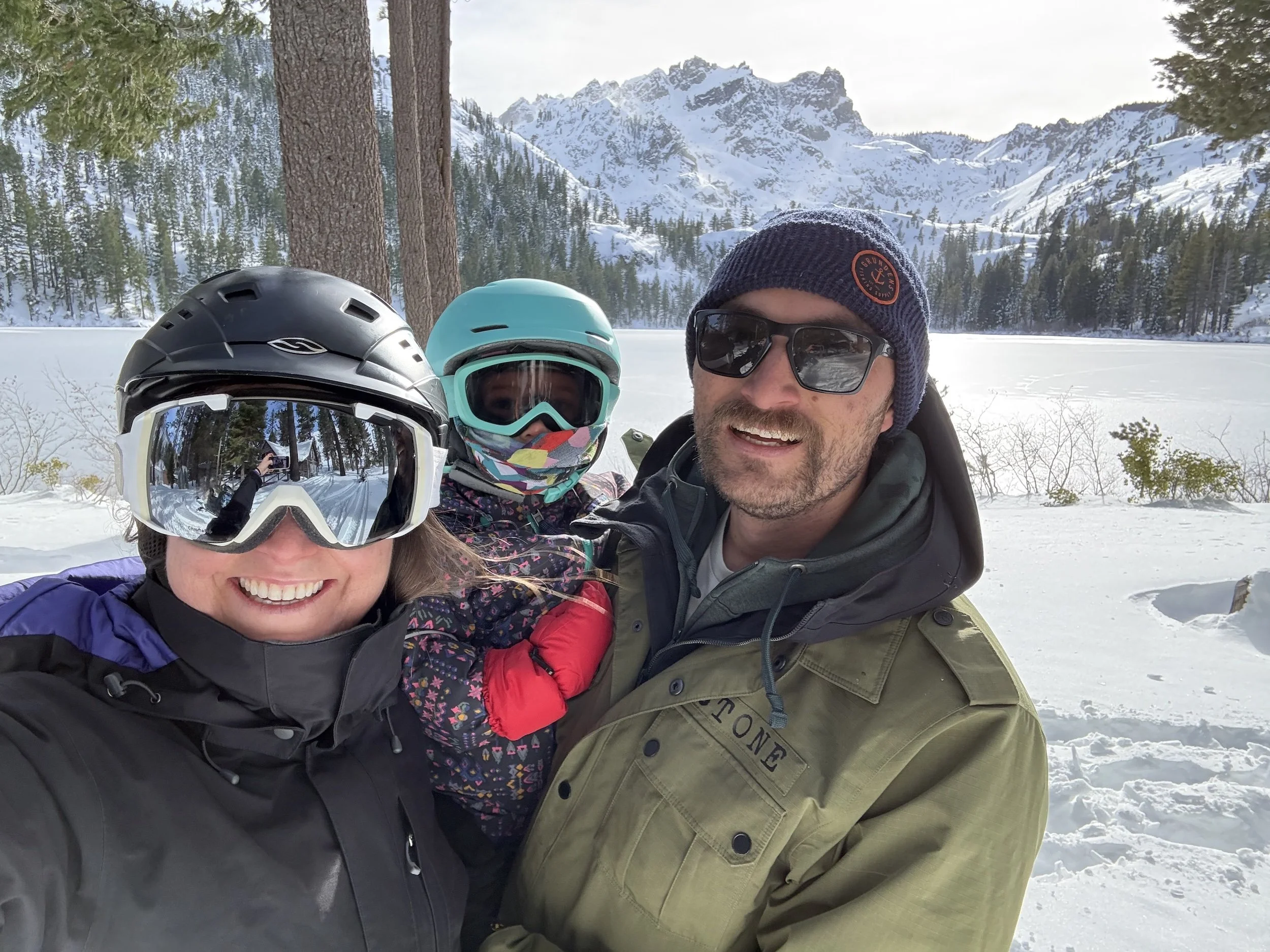 Group of three people taking a selfie outdoors in a snowy landscape, with snow-covered mountains and trees in the background. One person is wearing a black helmet and ski goggles, another a turquoise helmet and sunglasses, and the third is wearing a navy beanie and glasses.
