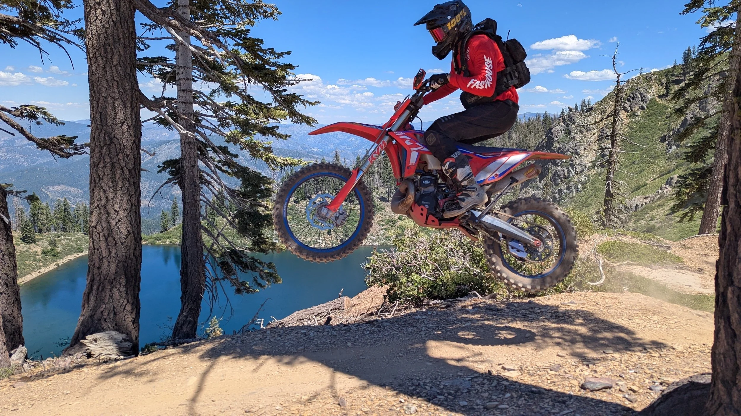 A person riding a red dirt bike off-road through a forested mountainous area with a lake and distant mountains in the background.