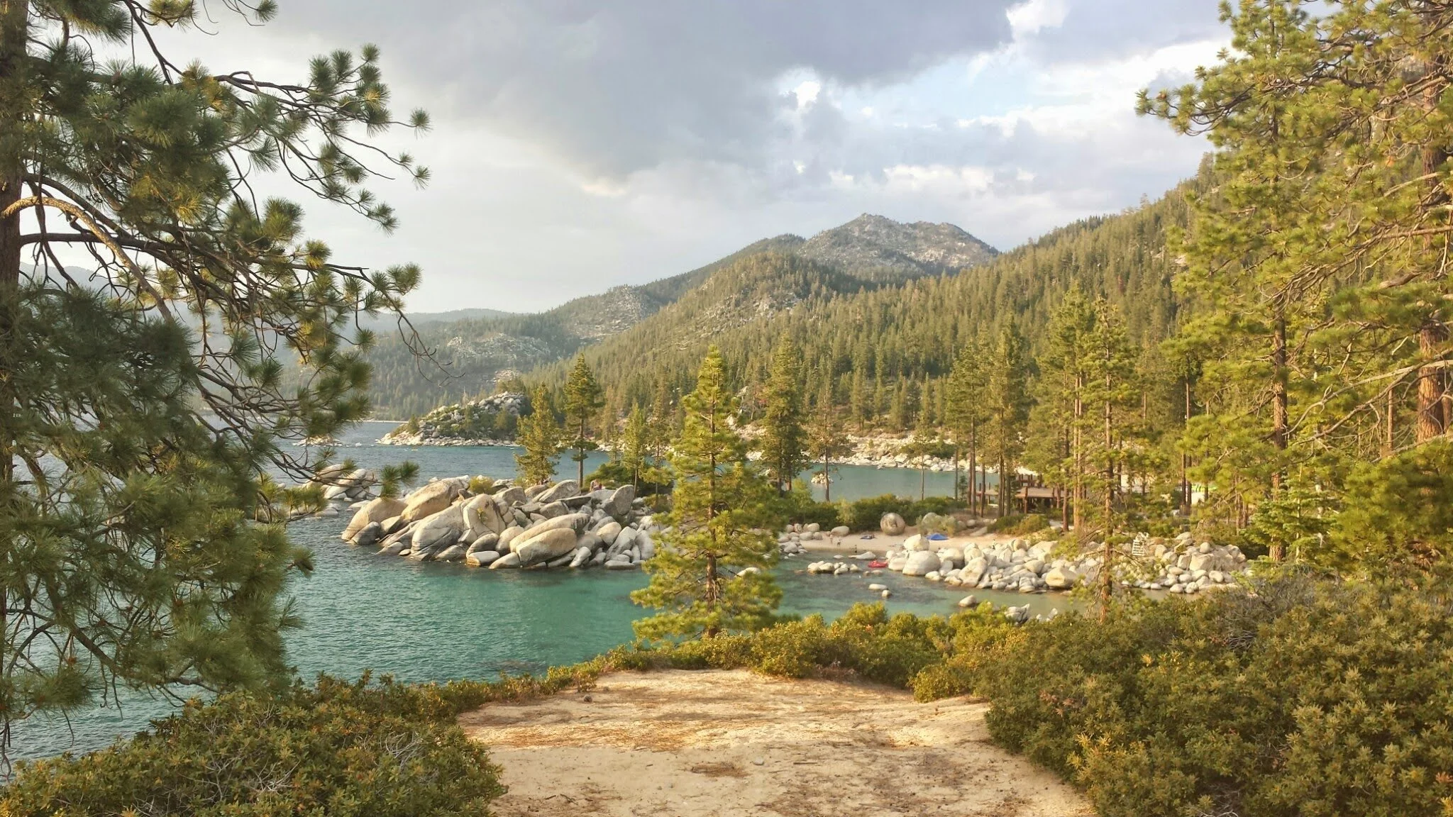A scenic mountain landscape on East Lake Tahoe with a lake, large rocks along the shoreline, and dense green pine trees under cloudy sky.