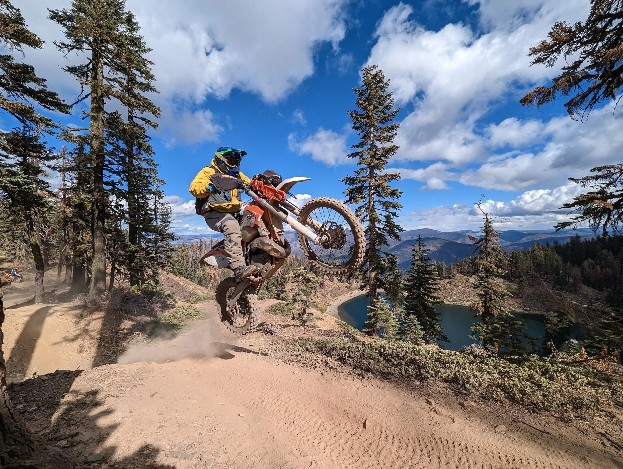 Cooper riding a dirt bike in mid-air during a jump on a mountain trail surrounded by trees and scenic Northern Nevada mountains in the background.