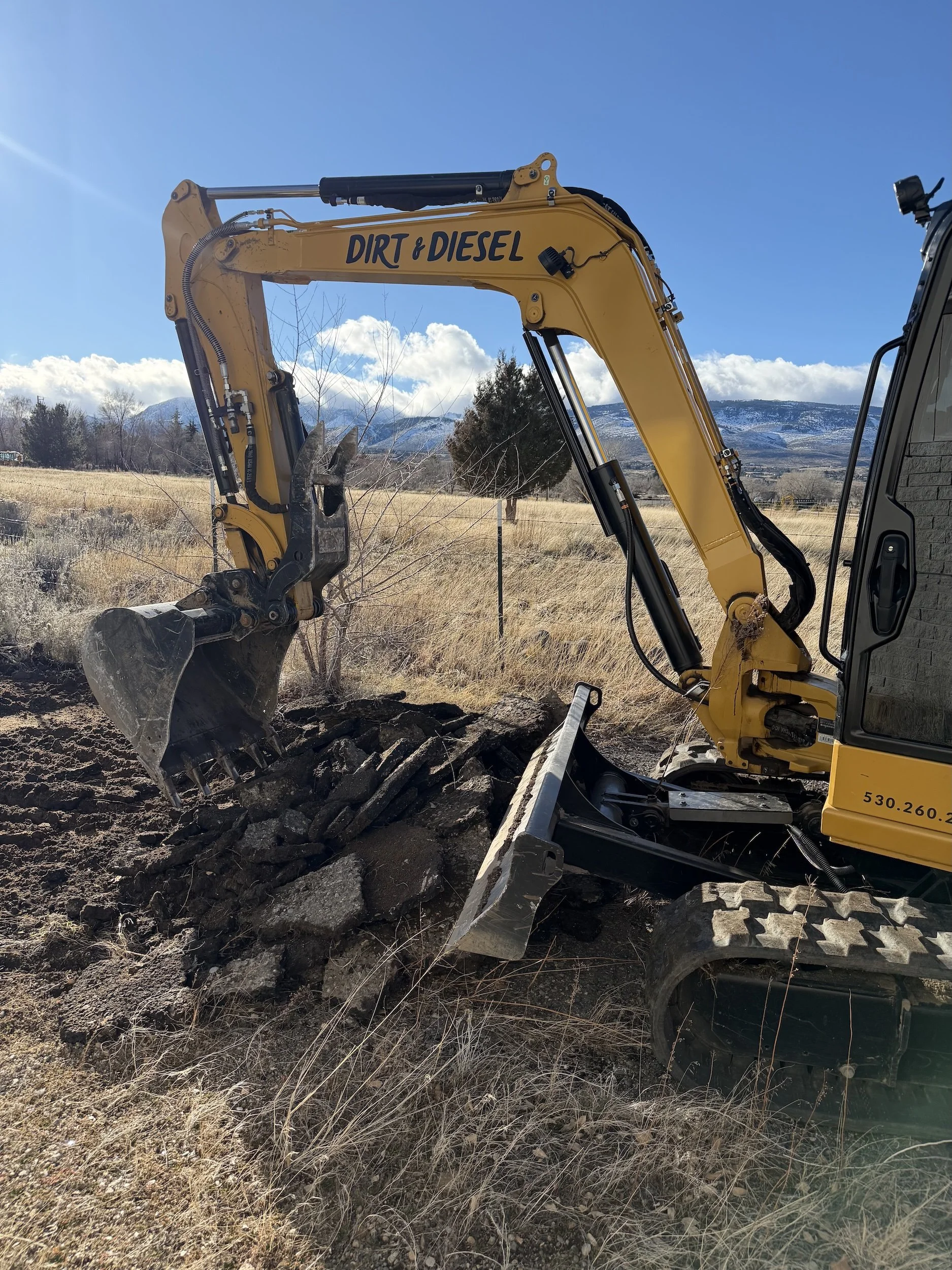 A yellow excavator with a Dirt & Diesel decal parked in a dry grassy field with a fence line and mountains behind.