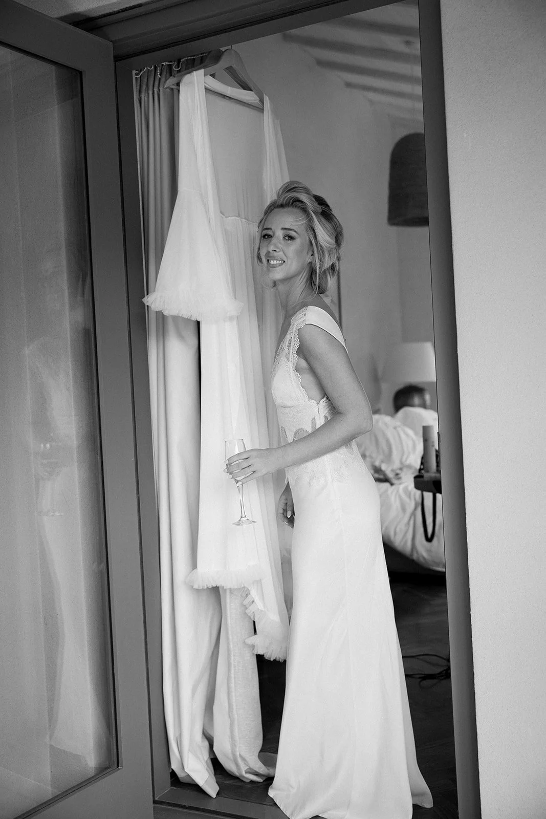 A woman in a wedding dress holding a glass of champagne, standing by a closet with a gown hanging, in a room with a man in bed in the background.