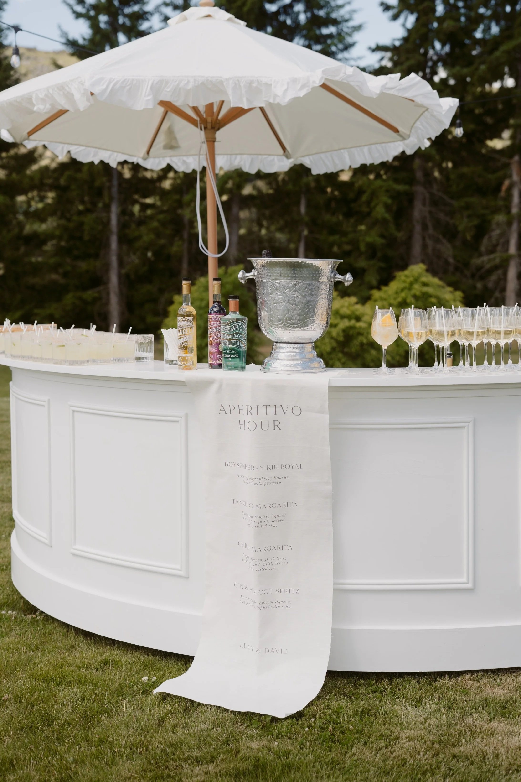 Outdoor white bar with a large umbrella providing shade, decorated with bottles of alcohol, glasses of cocktails, and a menu banner, set on a grassy area with trees in the background.