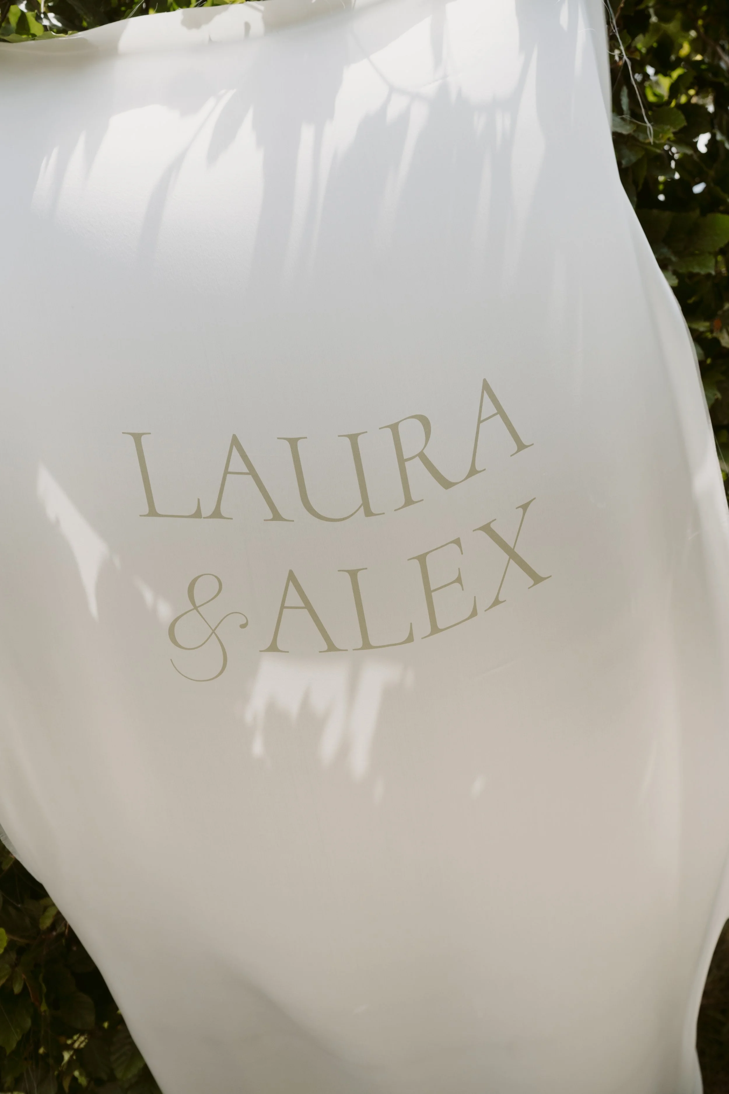 A white paper or bag with the words 'Laura & Alex' printed on it, hanging outdoors with shadows of leaves cast on it.