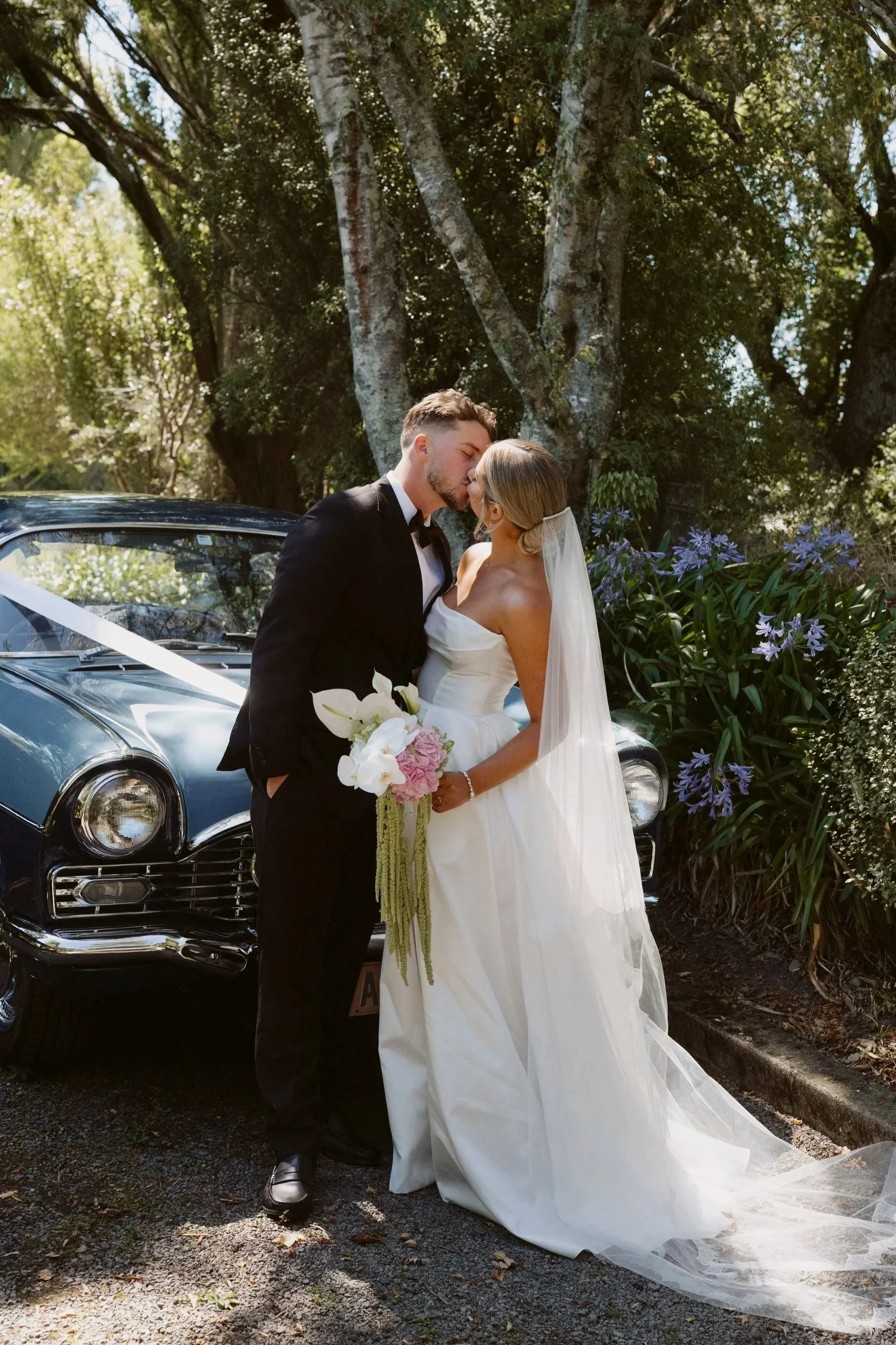 A newlywed couple sharing a kiss in front of a black vintage car outdoors, surrounded by large trees and flowering plants, during daytime.