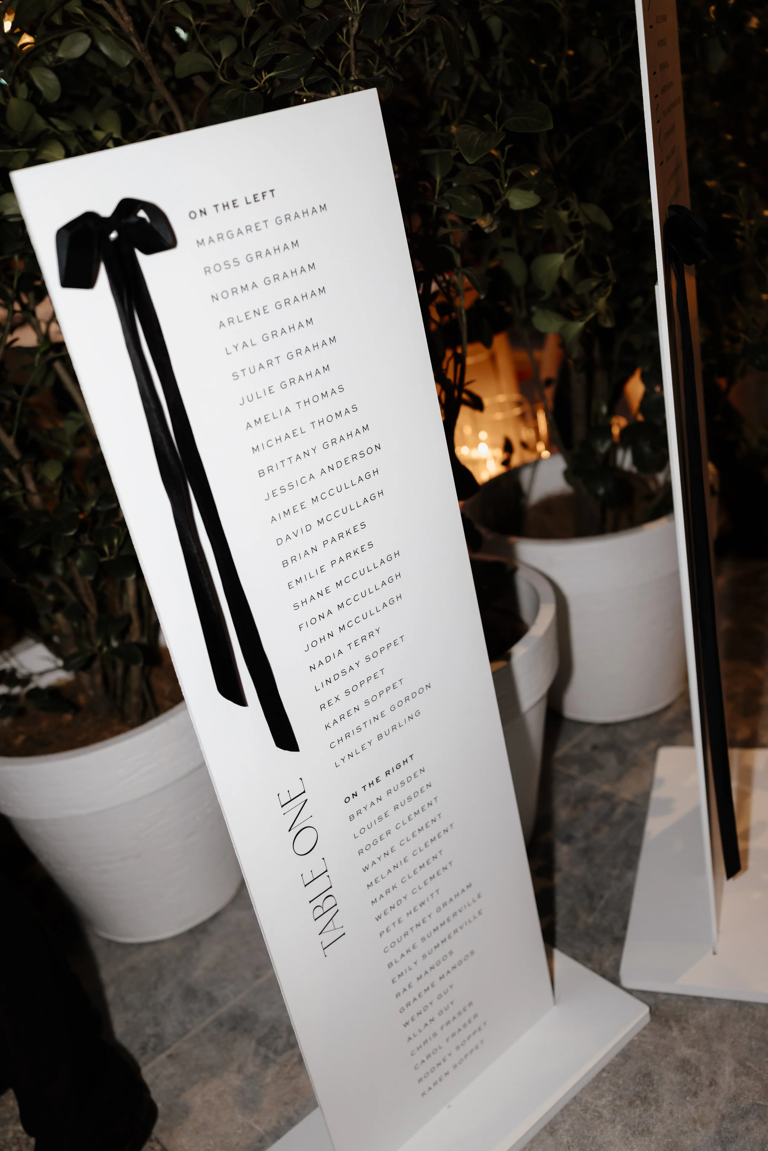 A white, freestanding signboard with a black ribbon illustration at the top, listing names of people involved in an event titled 'ON THE LEFT' and 'ON THE RIGHT' in black text. The sign is positioned outdoors with green potted plants and a warmly lit background.