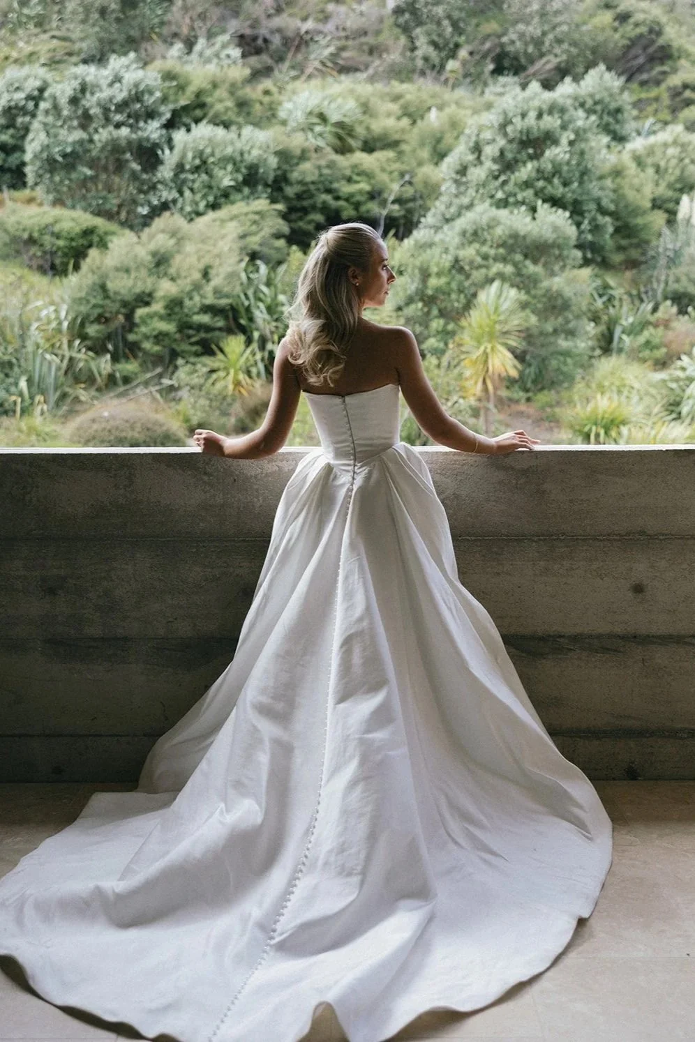 A woman in a strapless white wedding gown stands on a balcony, looking out at lush green trees and foliage in the background.
