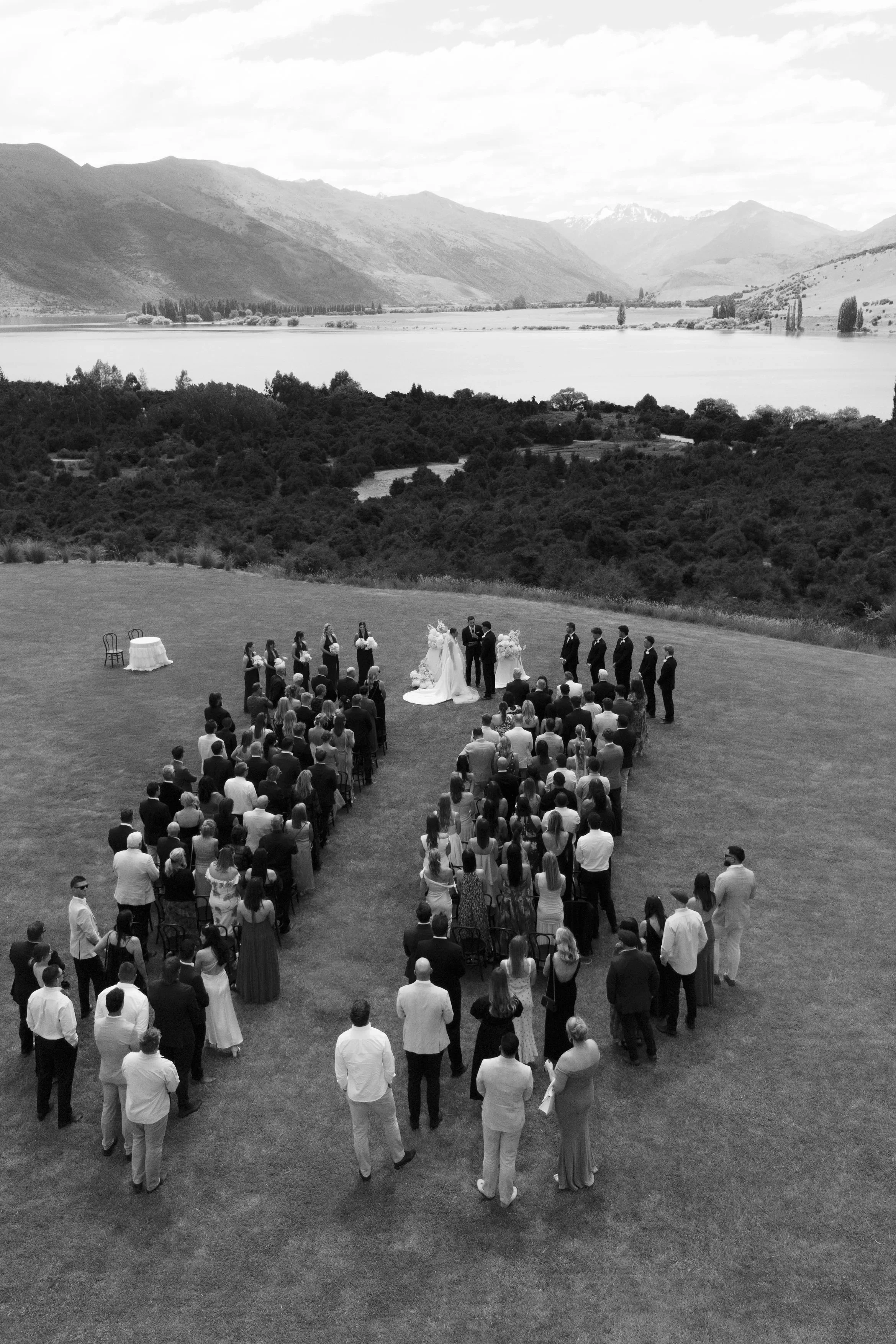 Black and white photograph of a wedding ceremony outdoors near a lake with mountainous landscape in the background. Guests sit and stand on a grassy area, facing the officiant and couple, who are standing under a floral arch.