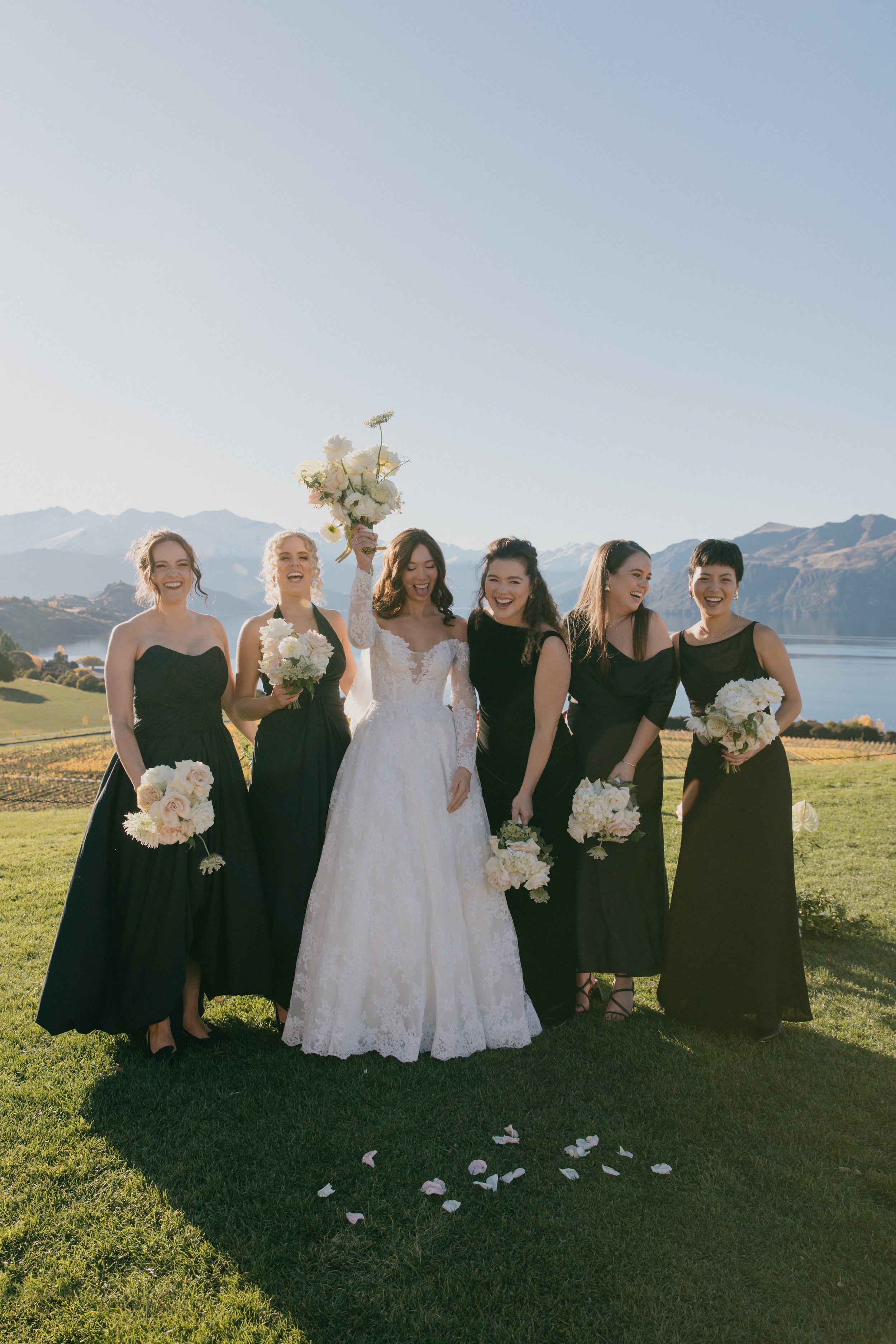 A bride in a white lace wedding dress holding a bouquet, standing among five women in black dresses, outdoors on a grassy field with mountains in the background, during daytime.