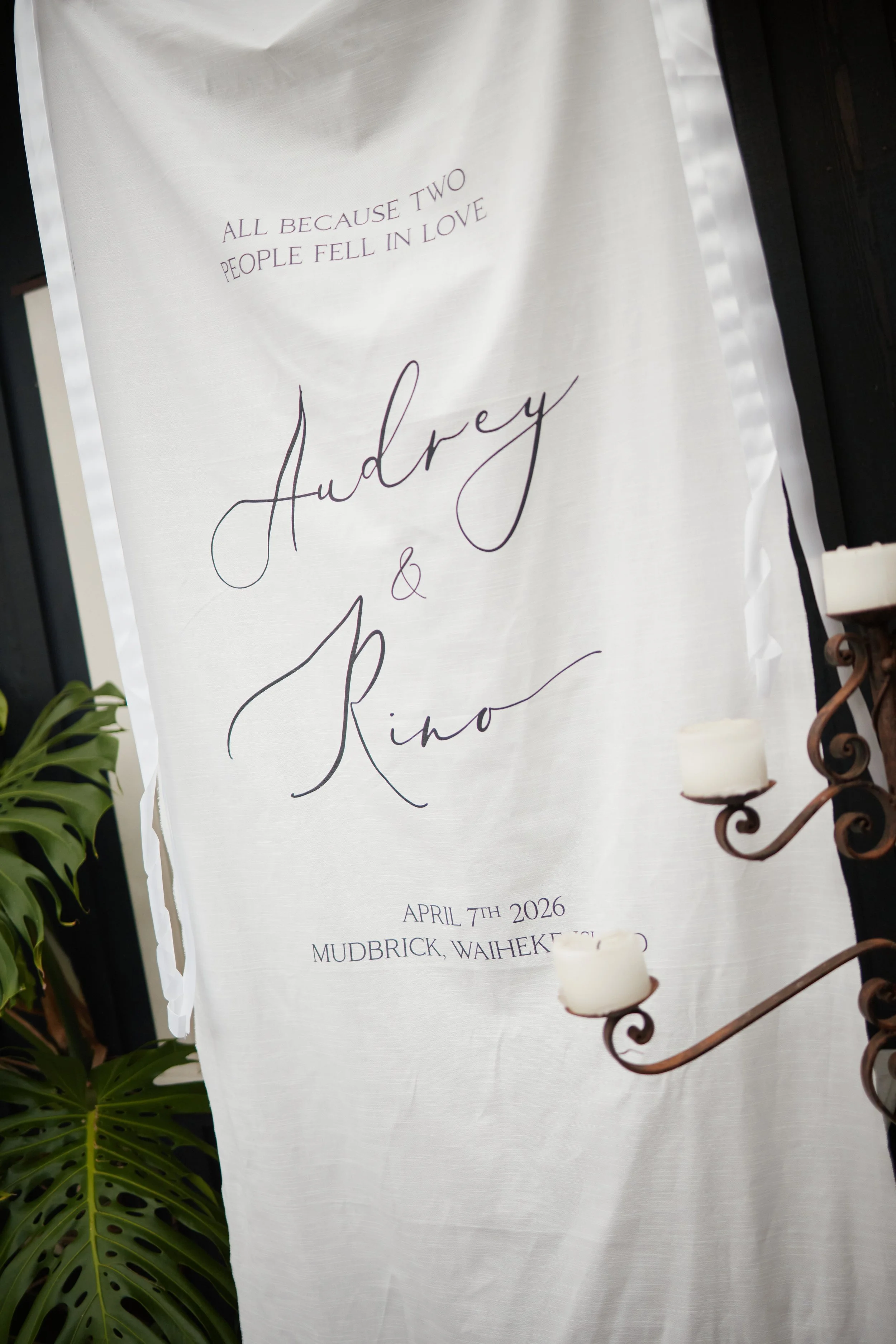 Wedding invitation banner with elegant script text, reading 'All because two people fell in love, Audrey & Rino, April 7th 2026, Mudbrick, Waiheke,' on a white background with greenery on the side and candles in front.
