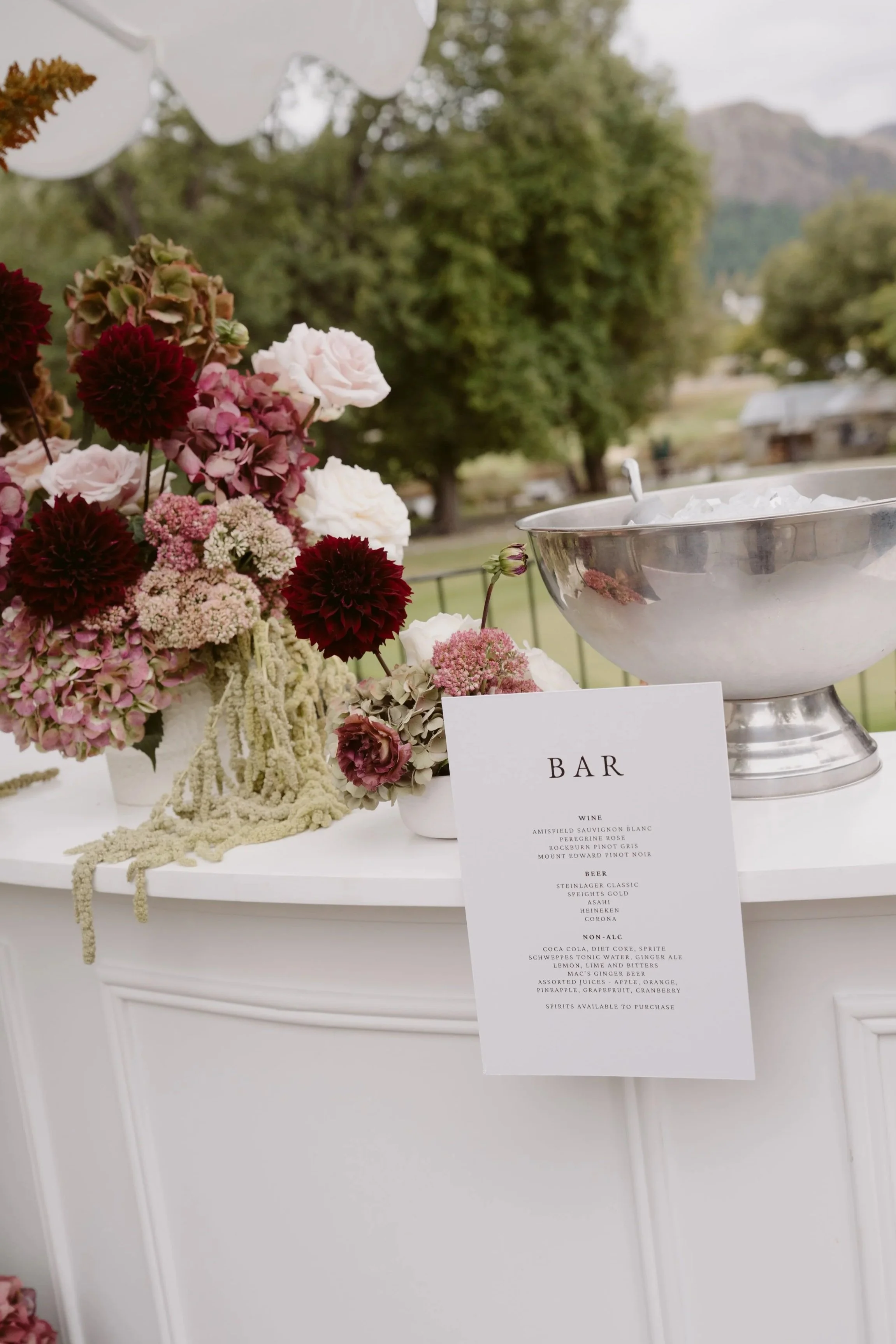 A floral arrangement with pink, white, and dark red flowers on a white table, with a silver bowl labeled "bar" containing ice, and a menu listing wine, beer, and non-alcoholic drinks, outdoors with trees and mountains in the background.