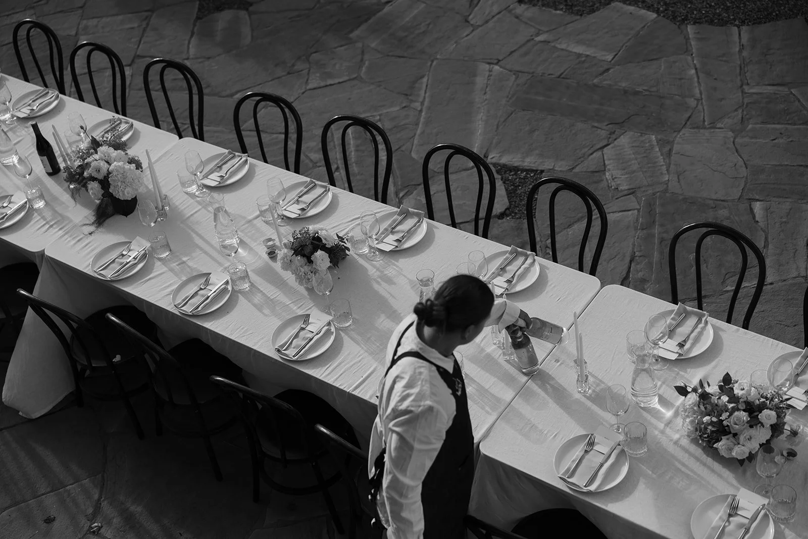 A long dining table set with plates, forks, knives, glasses, and candles, decorated with floral centerpieces, in a stone-floored room, with a woman cleaning a bottle on the table.