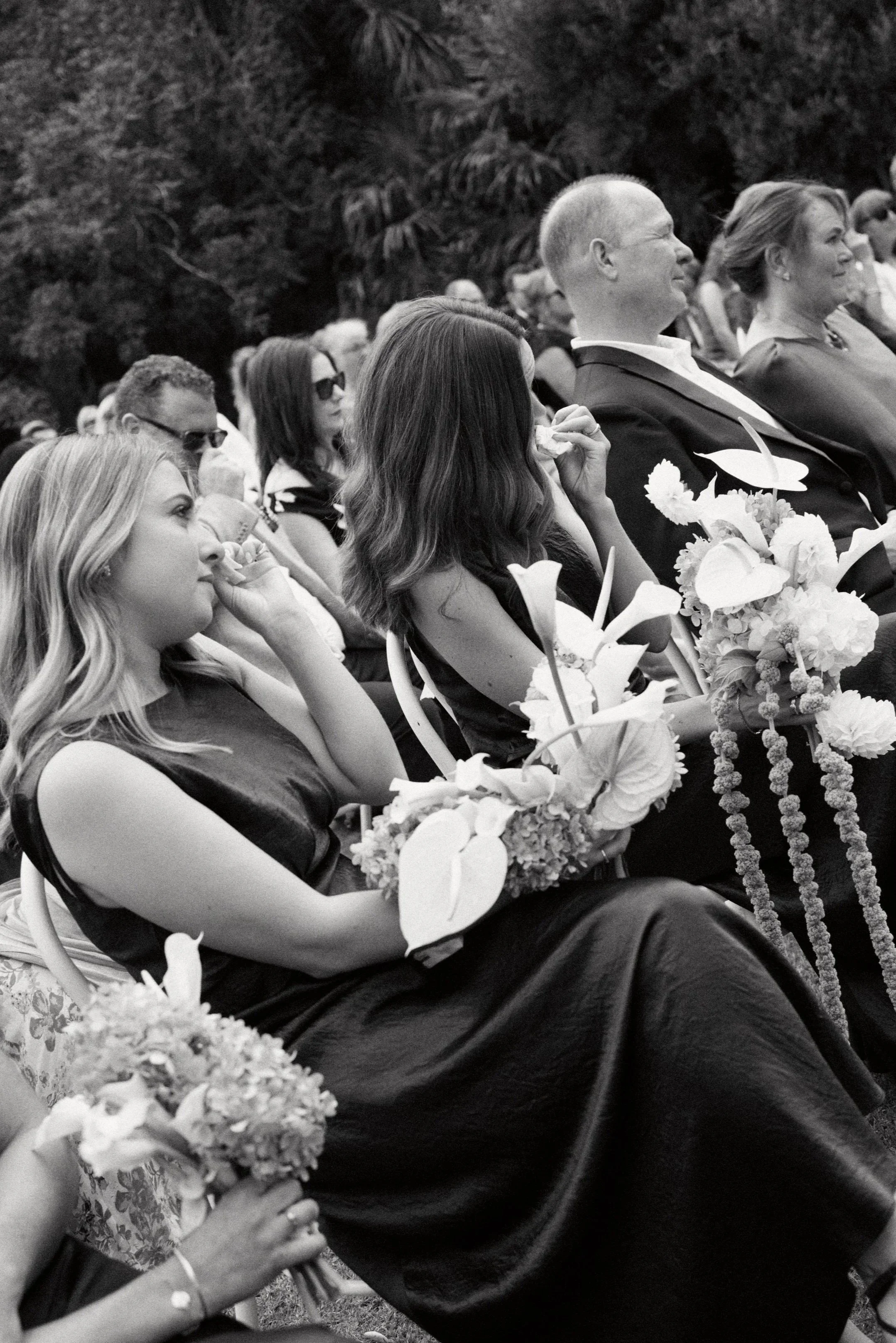 A group of people attending an outdoor event, sitting on chairs, with some holding bouquets of flowers, and trees visible in the background.