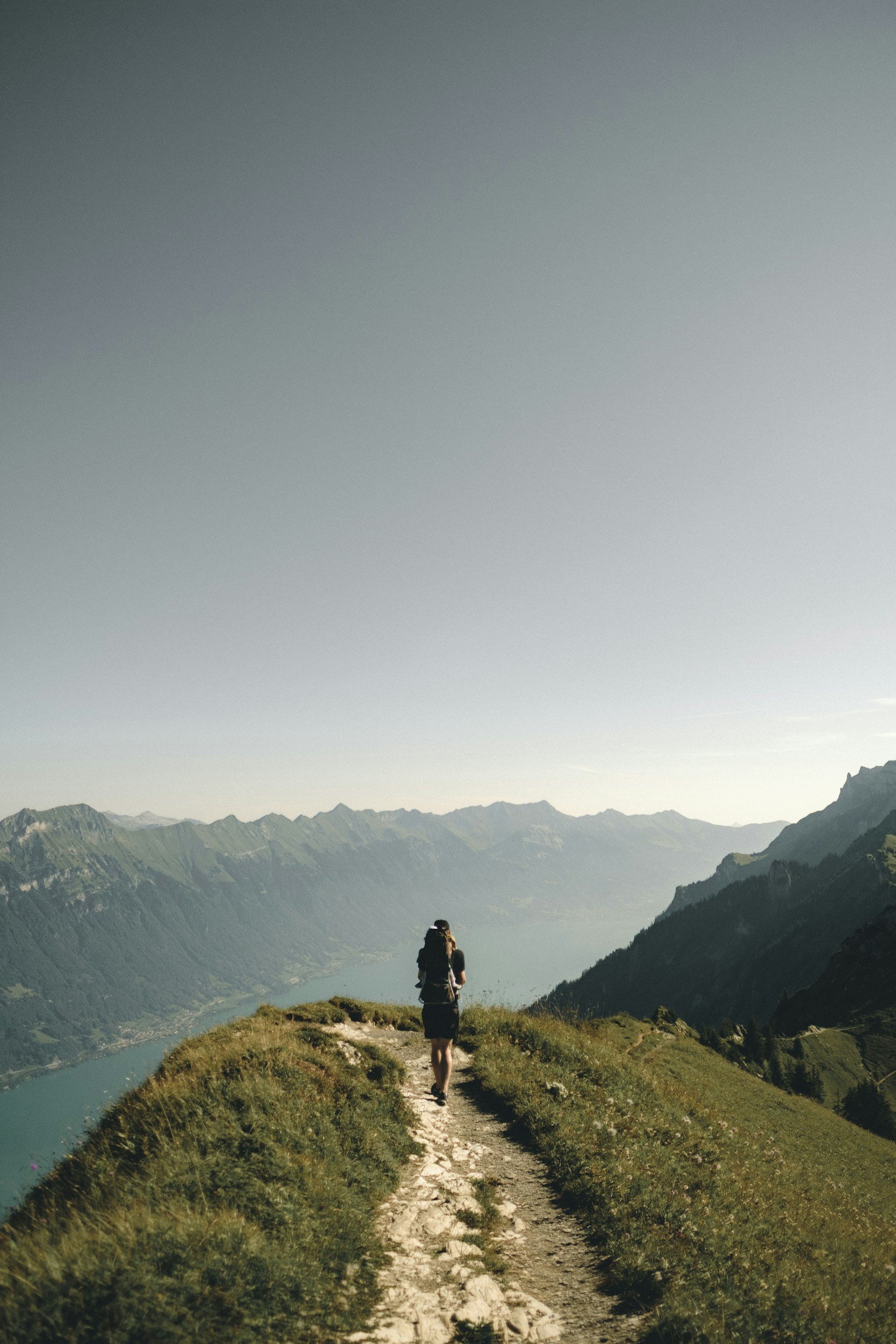 A person hiking uphill on a narrow dirt trail through green grass with mountains and a lake in the background.