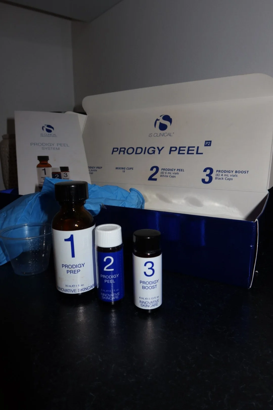 Set of skincare products, including Prodigy Peel system bottles labeled 1, 2, and 3, on a black surface with a blue box in the background.