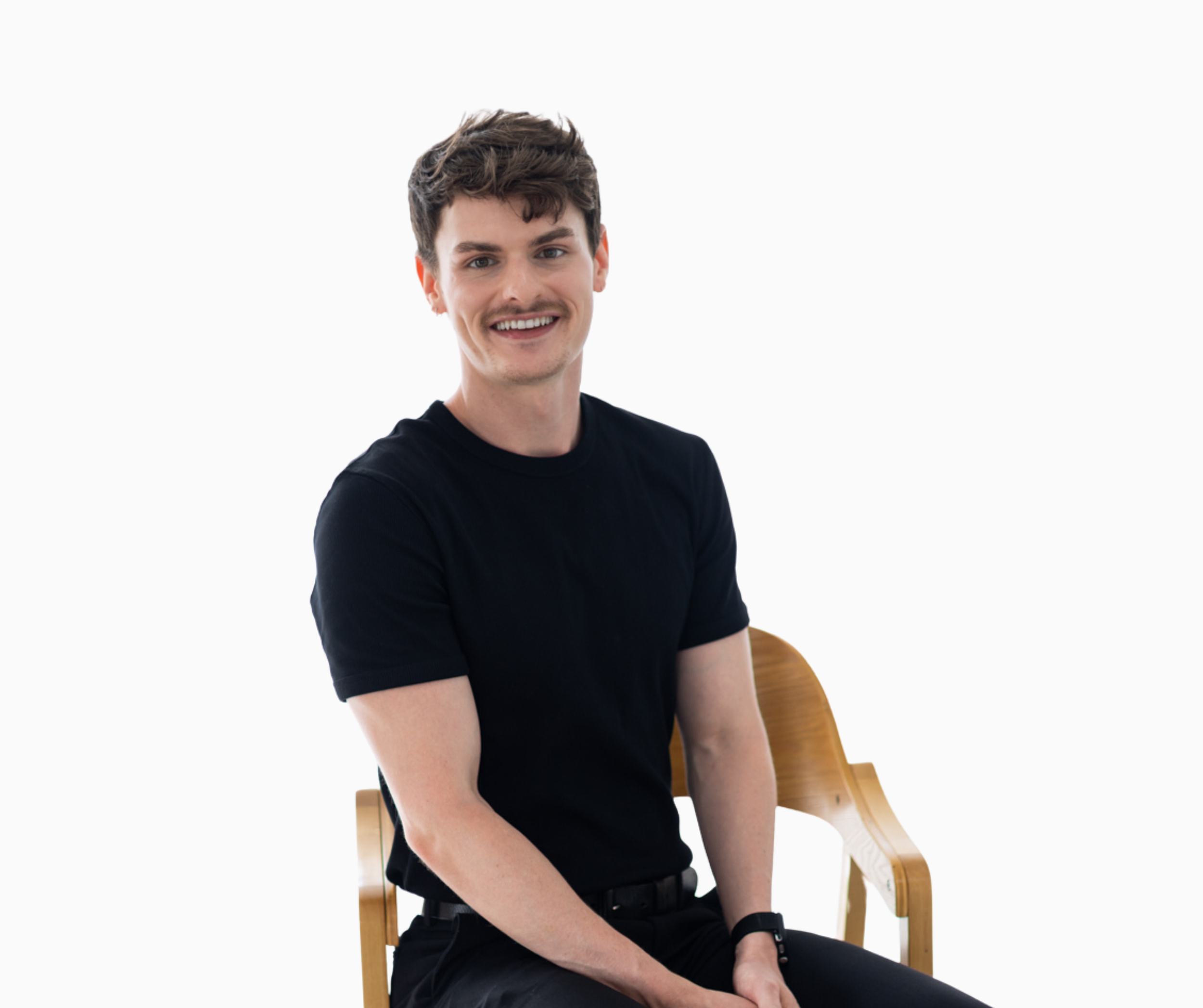Young man with short dark hair, mustache, and earrings, smiling, wearing a black patterned polo shirt, sitting on a wooden chair against a plain white background.