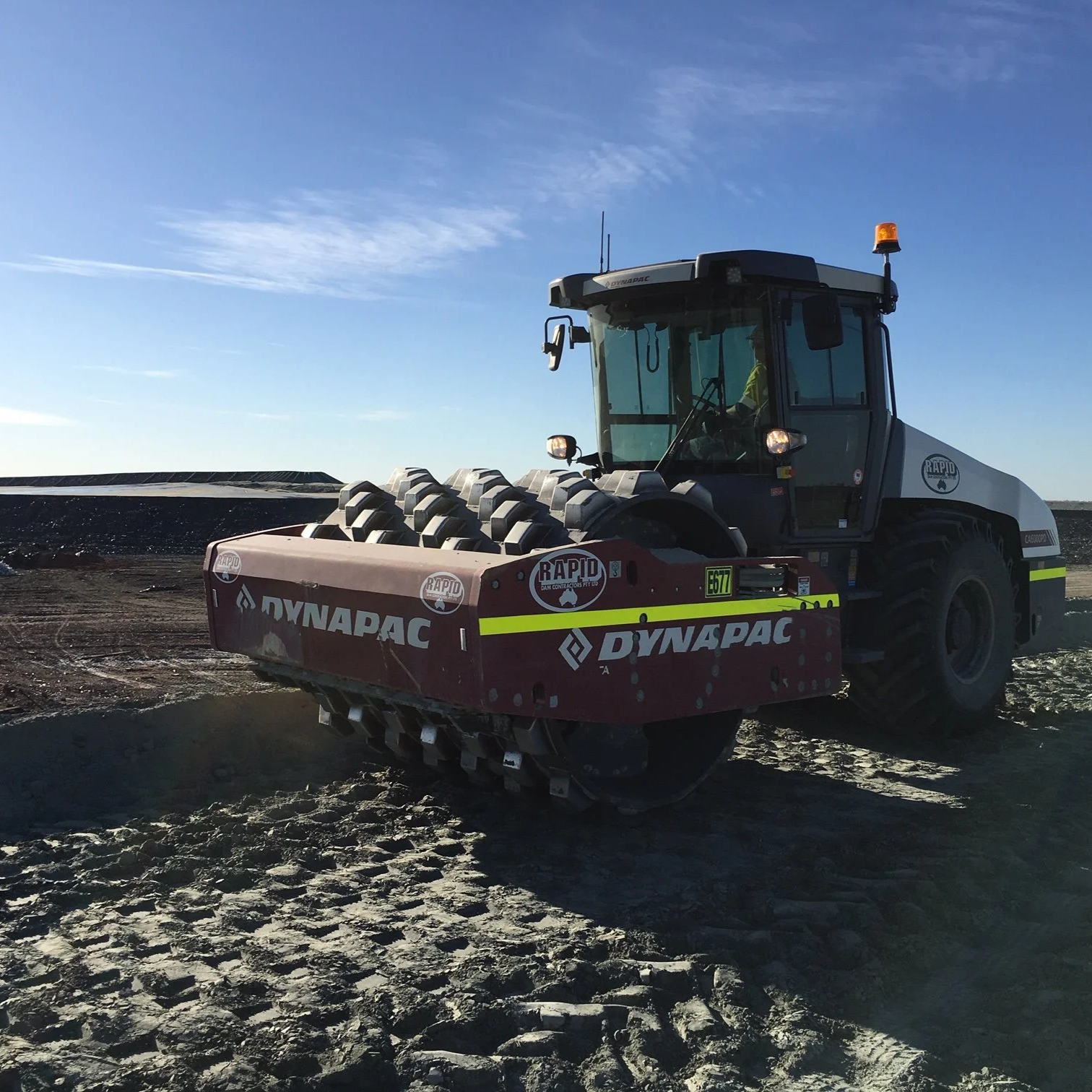 A Dynapac roller compactor on rocky terrain with a clear blue sky in the background.